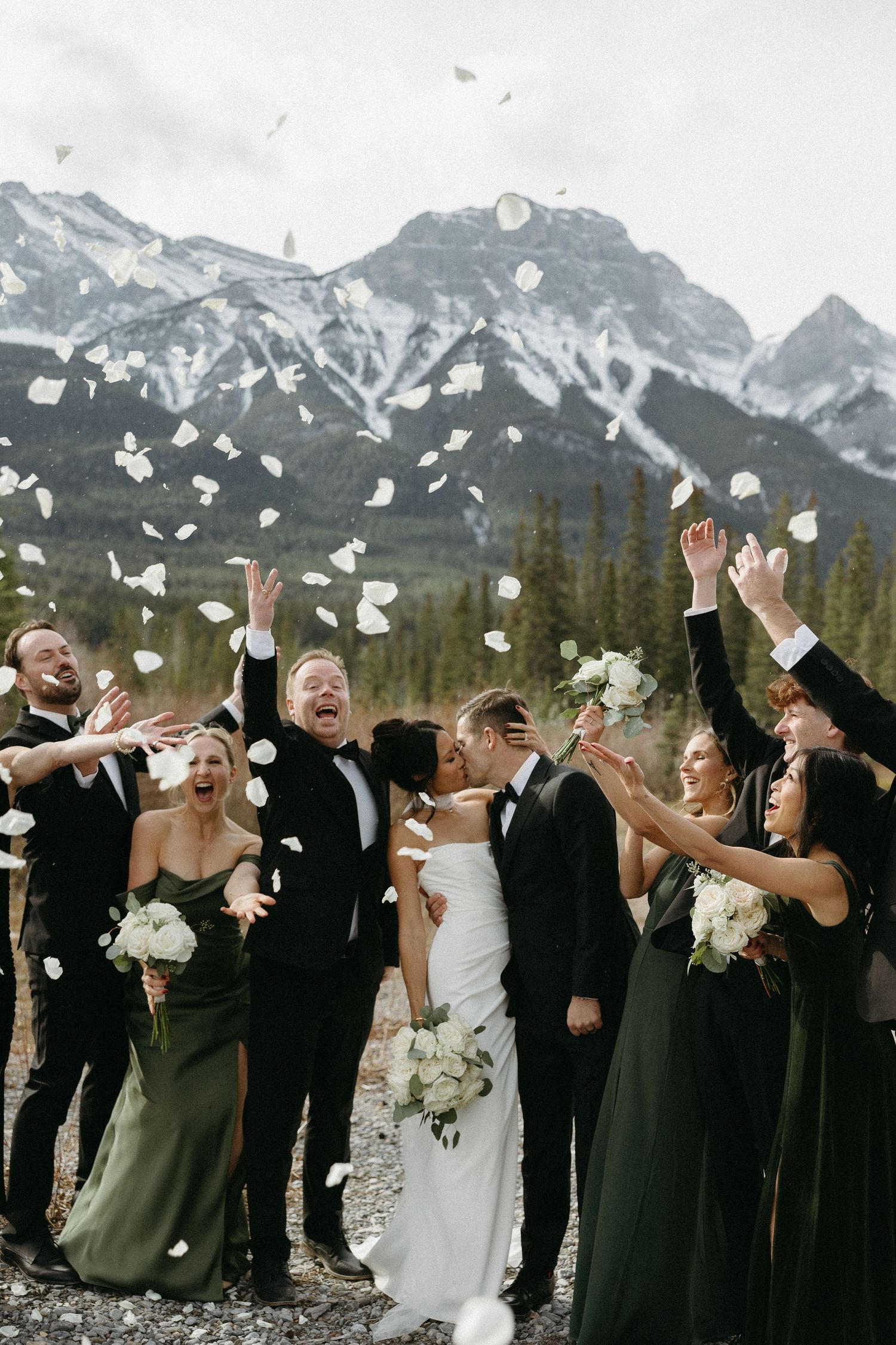 Wedding party celebrating with white petals tossed in air, bride and groom kissing, snow-capped mountains backdrop.