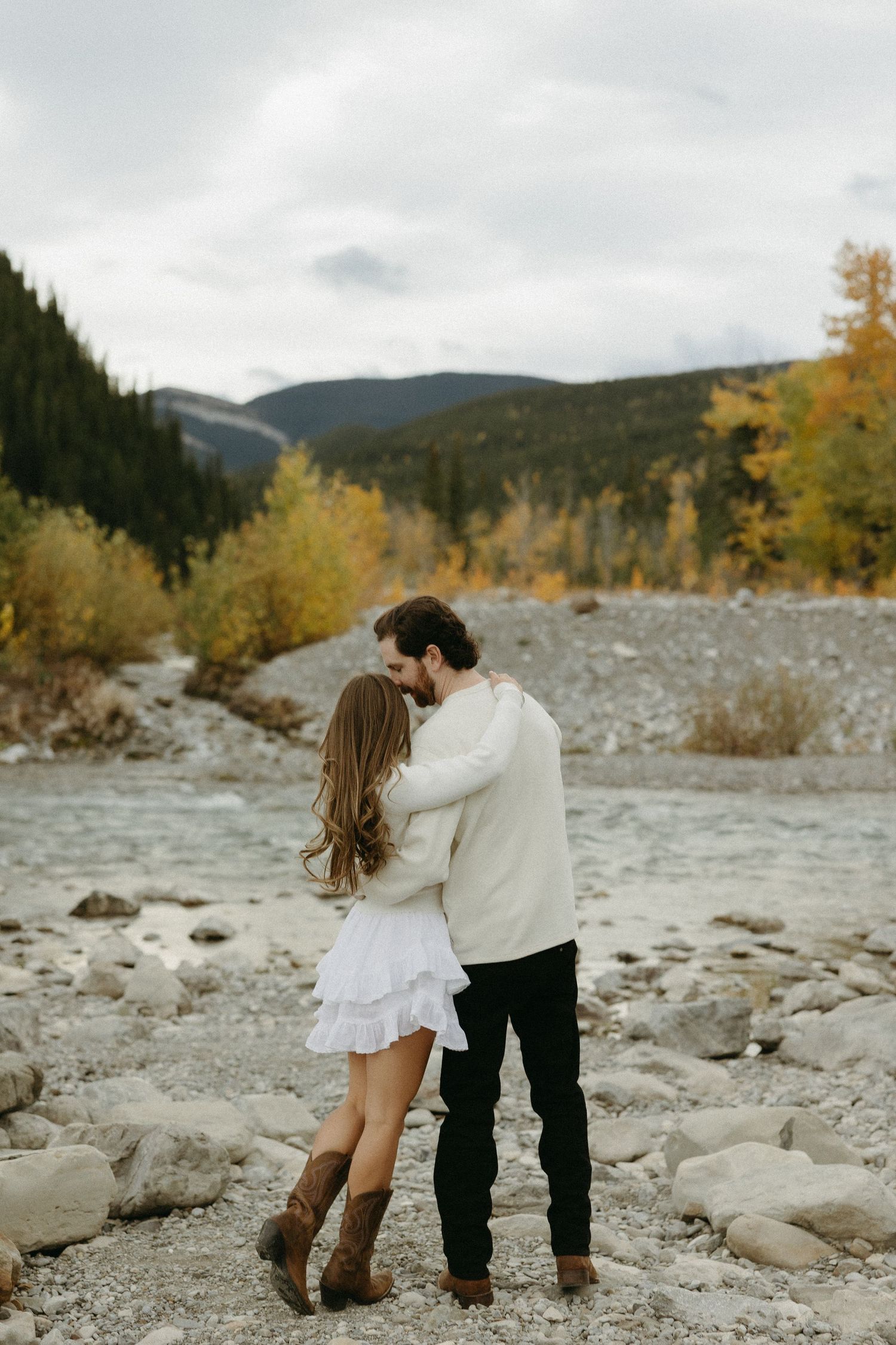 Couple kissing by a rocky river with autumn trees and mountains in the background during a fall engagement session.