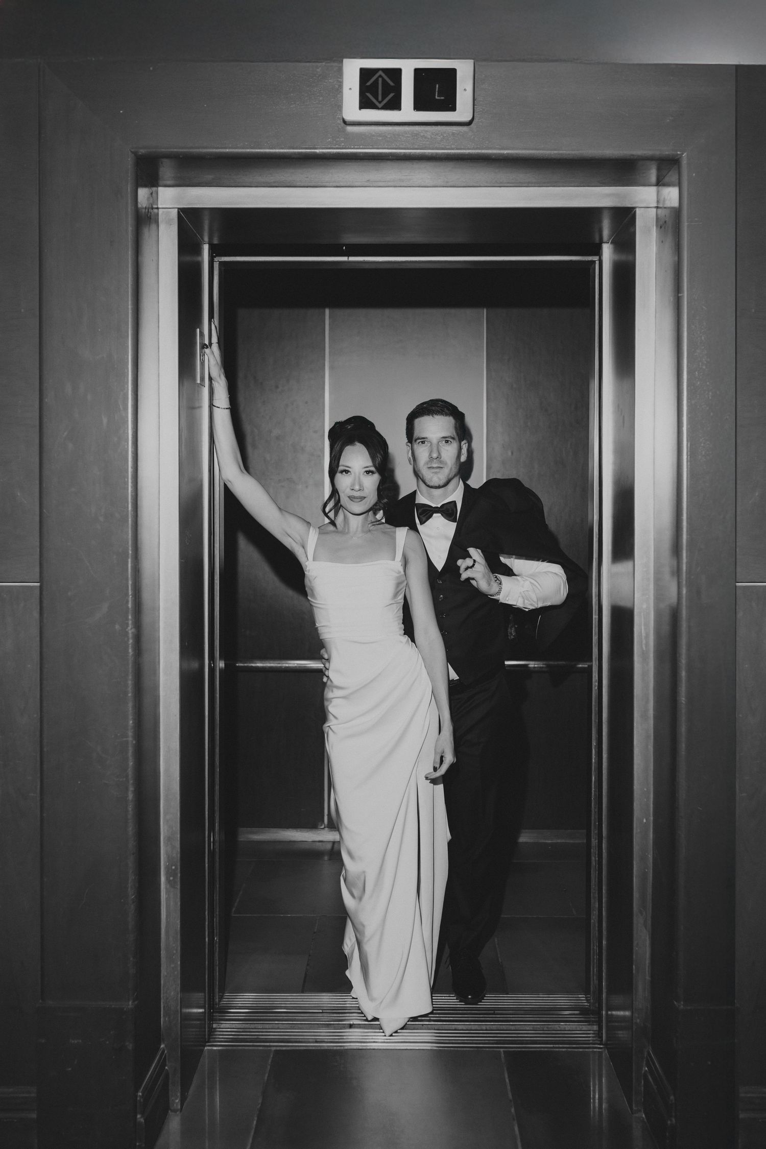 Wedding couple making playful entrance through elevator doors in black and white.