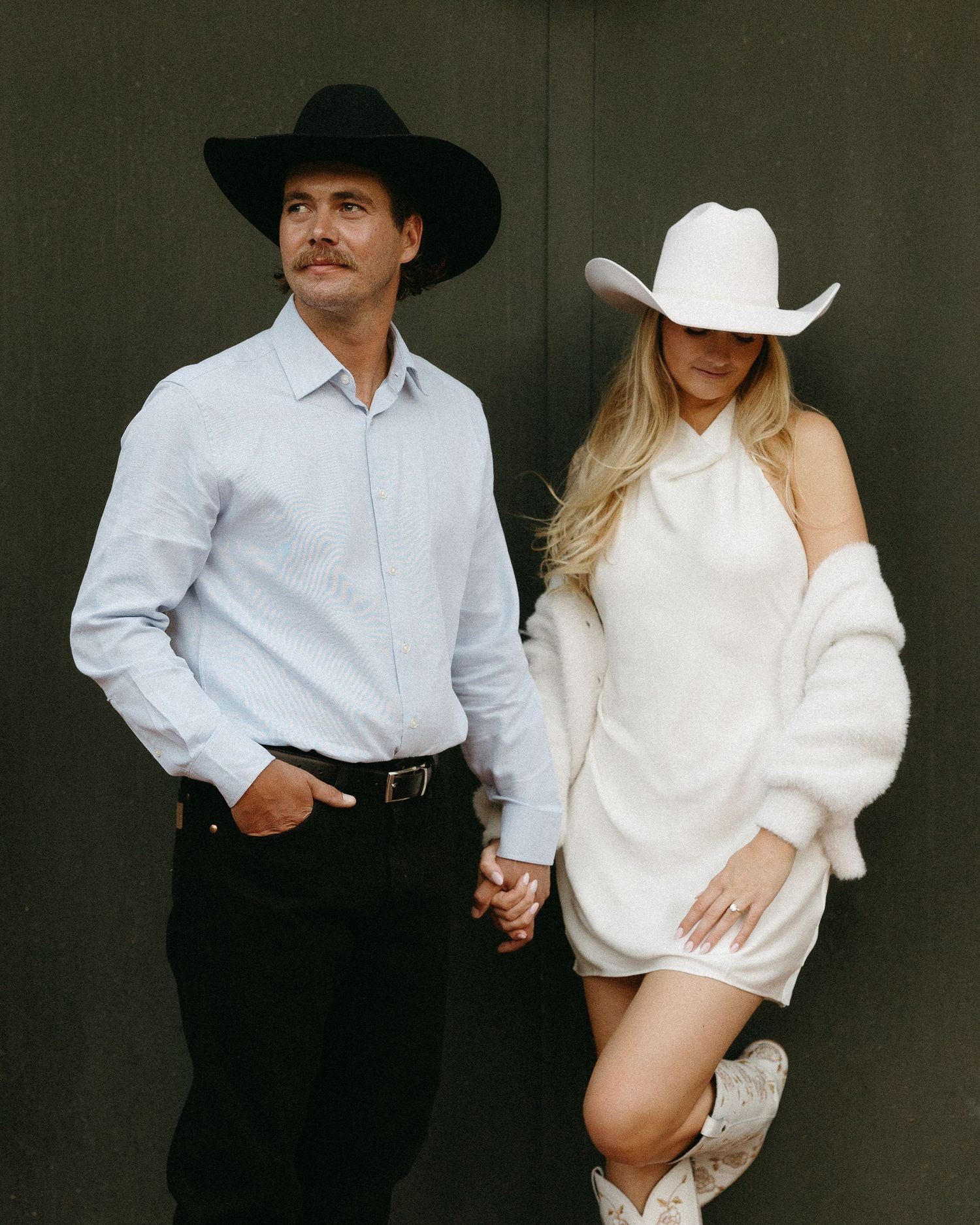 Stylish couple in contrasting black and white outfits pose against dark background with cowboy hats.
