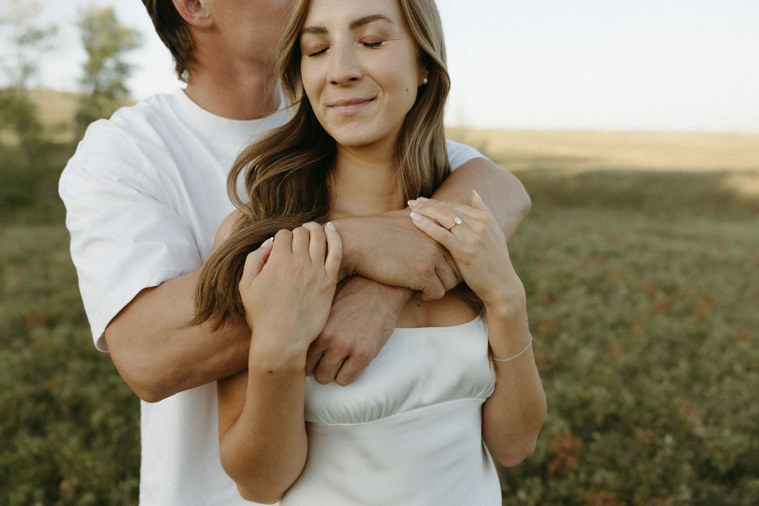 Romantic couple embraces in a sunlit field wearing white and casual clothing.