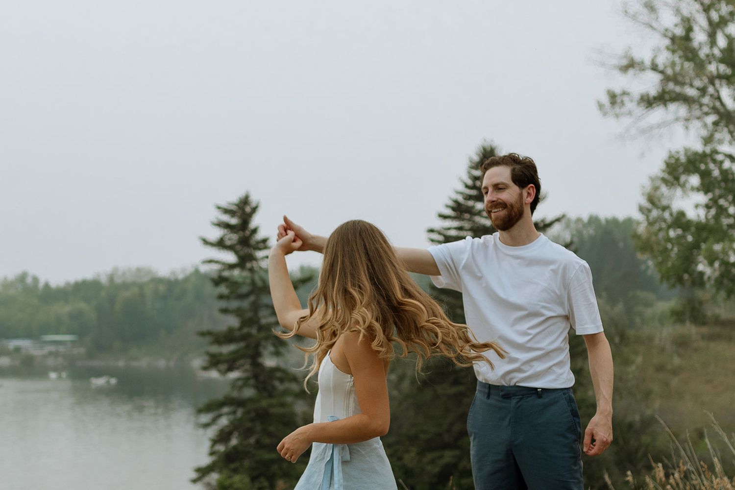 A couple in casual attire dances together on a grassy hillside with a lake in the background during sunset.