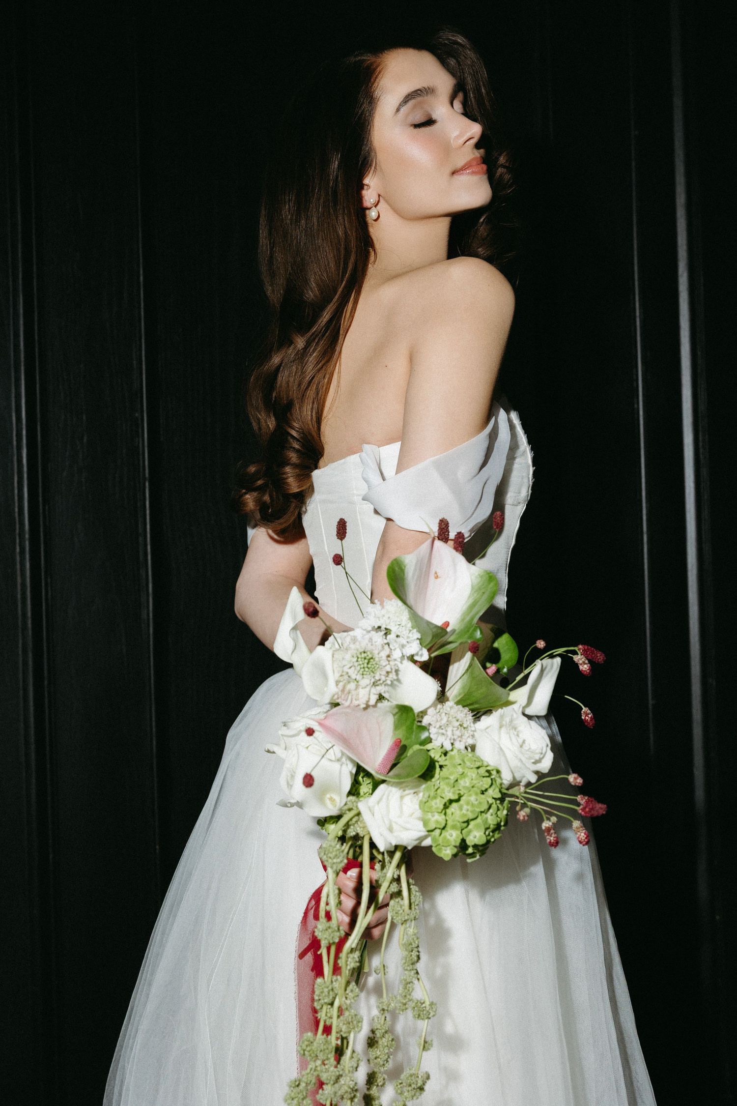 A bride in a white wedding dress holds a cascading bouquet of white flowers and greenery against a dark background.
