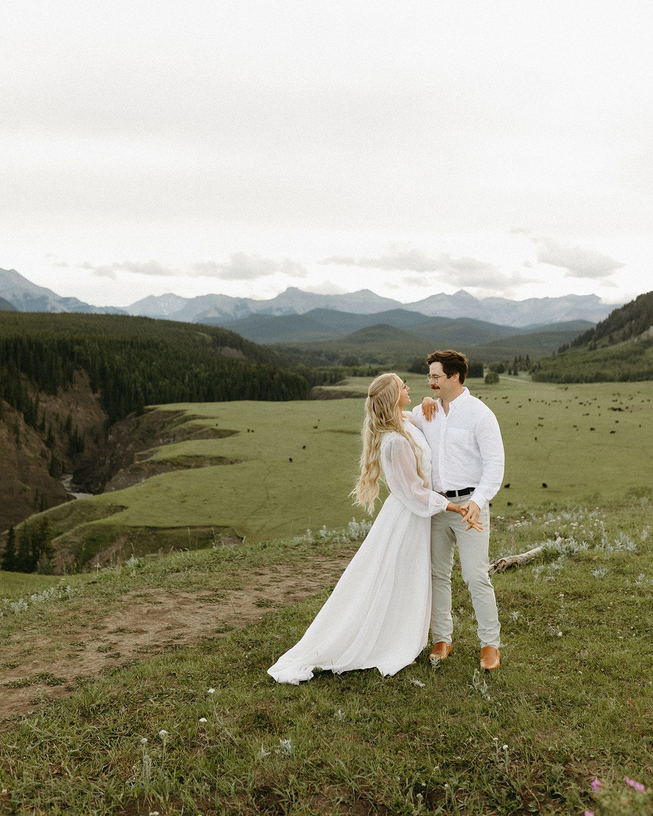 Couple in wedding attire standing on a scenic mountain meadow with sweeping valley and mountain range views.