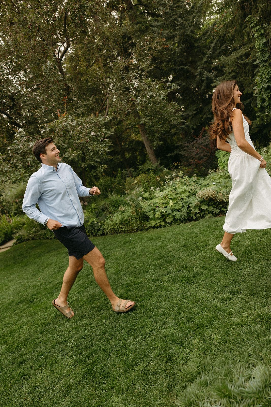 Playful couple runs and dances on a grassy hillside in casual attire.