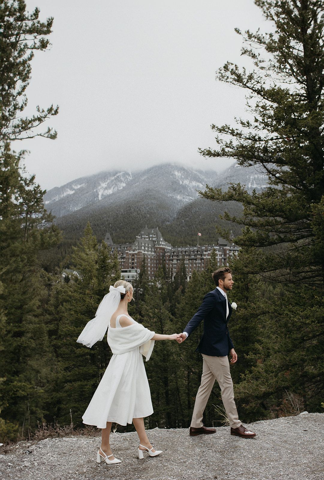 Wedding couple poses dramatically on mountain ledge with evergreen forest backdrop.