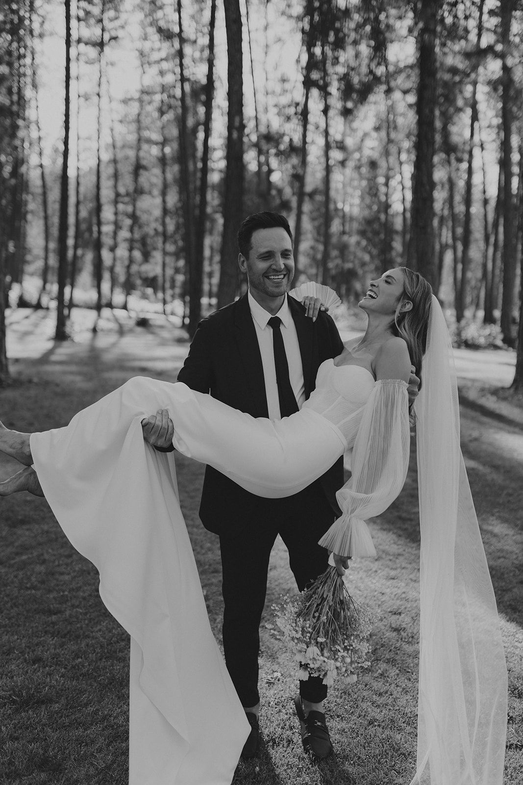 Joyful black and white wedding portraits among pine trees bride being carried by groom candid
