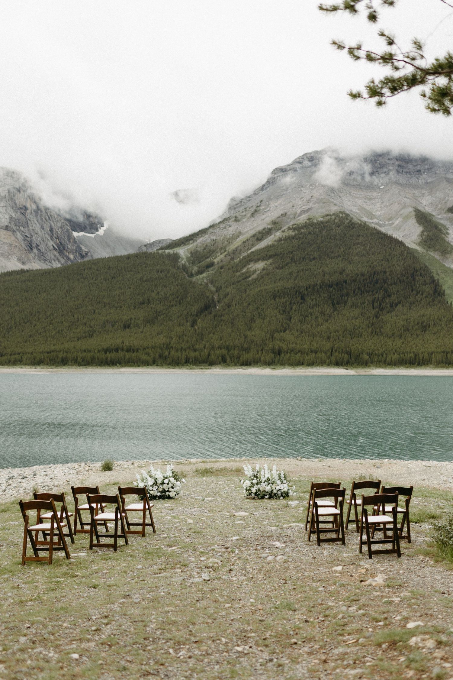 Outdoor lakeside wedding ceremony setup with wooden chairs and floral arrangements, framed by misty mountains and turquoise water.