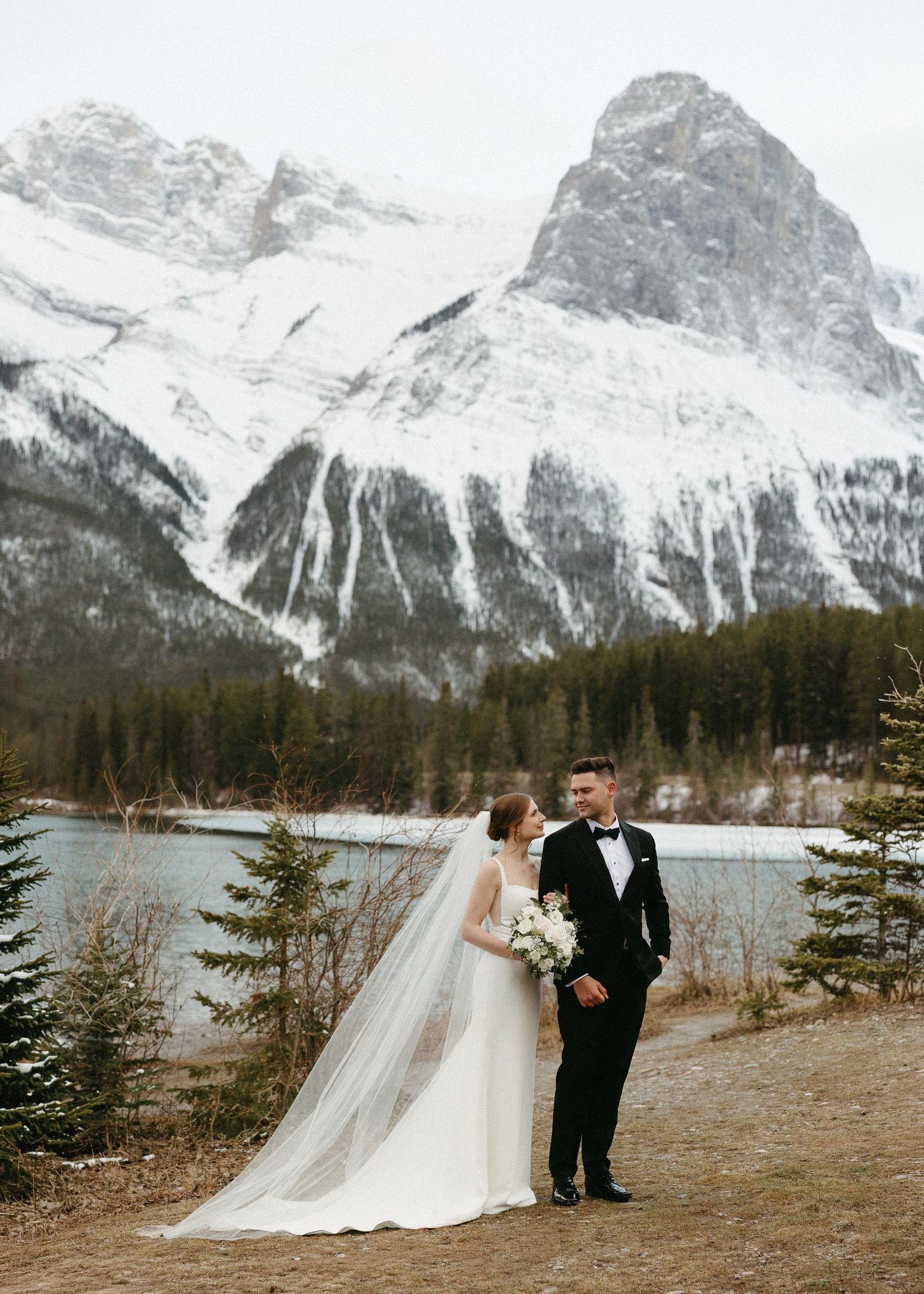 Bride and groom pose before snow-capped Rocky Mountains and turquoise lake in elegant winter wedding attire.