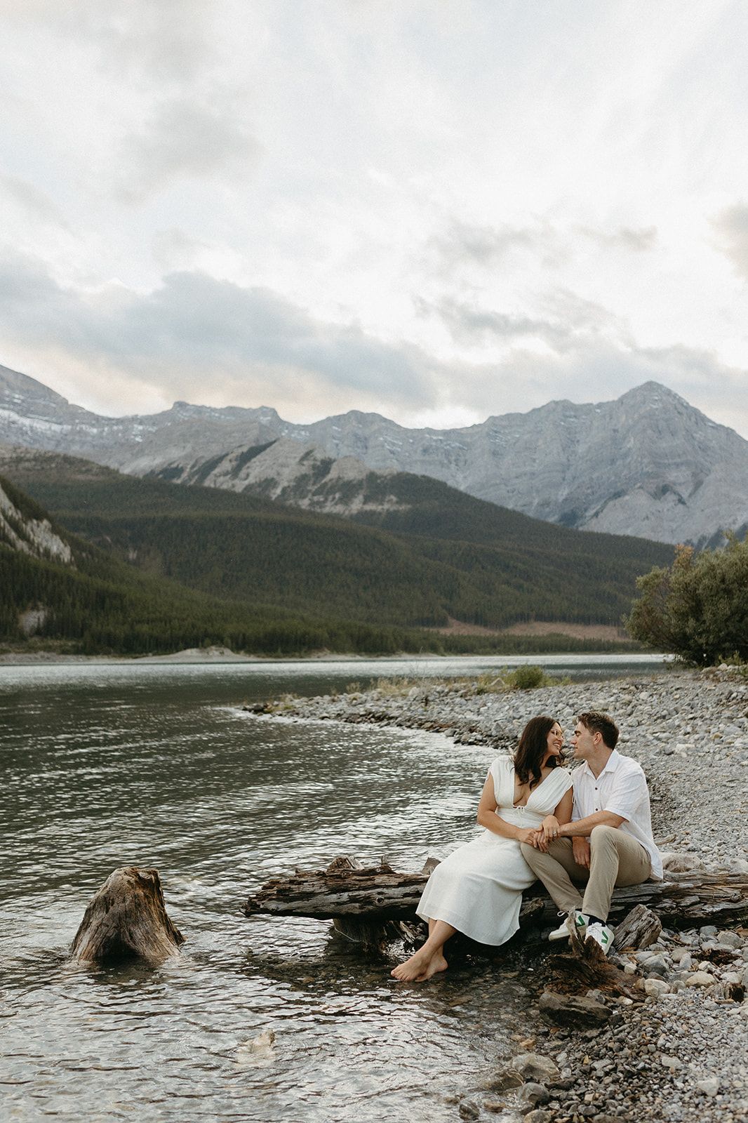 Couple sitting on rocks by a river with stunning mountain backdrop during an outdoor engagement session.