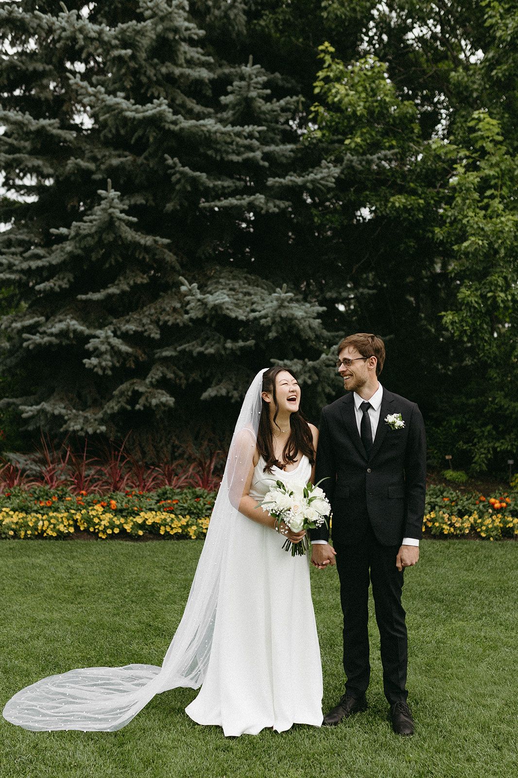 A bride and groom share candid moments in a garden setting with evergreen trees and colorful flowers in the background.