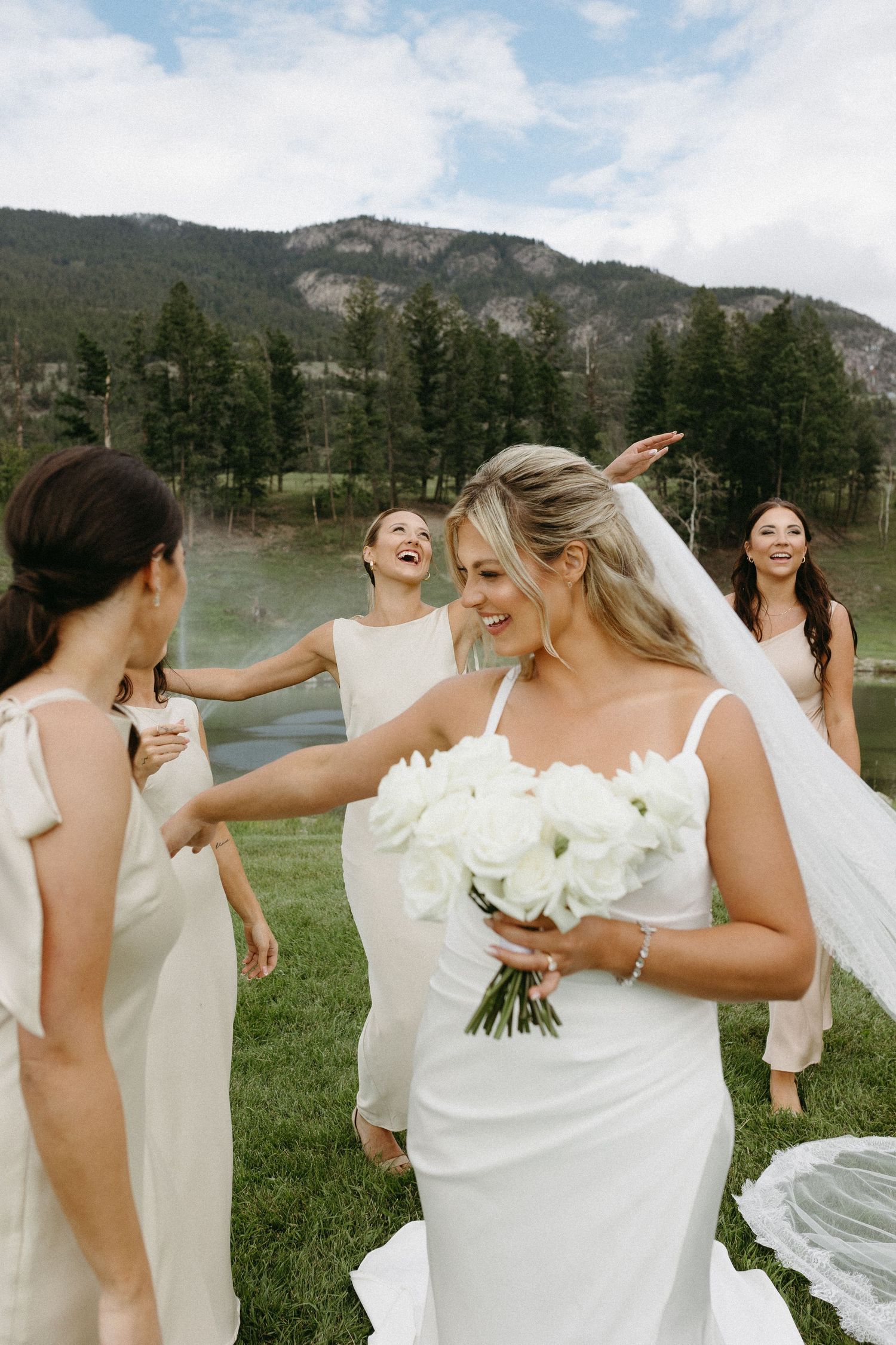 A joyful group in white dresses laughs together during an outdoor wedding celebration with mountains in the background.