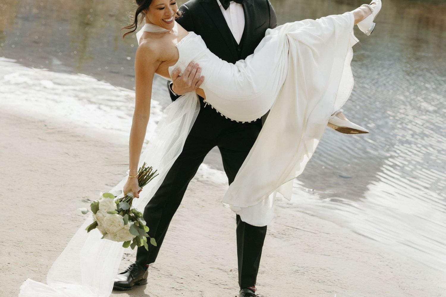 Romantic wedding couple poses by waterfront in formal attire.