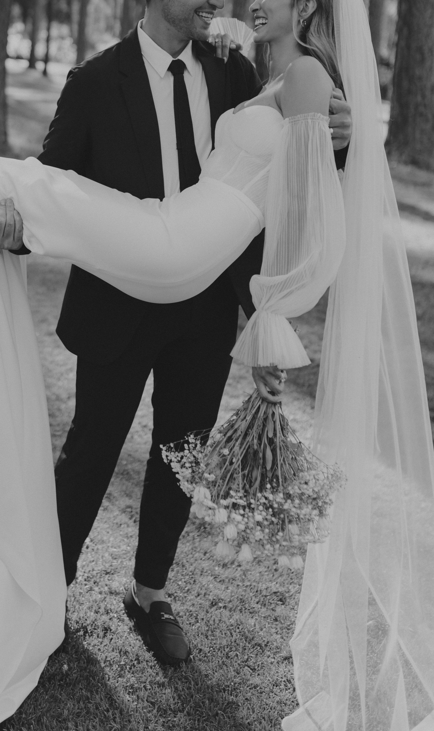 A black and white wedding photo showing a couple embracing outdoors, with the bride in a flowing dress holding dried flowers.