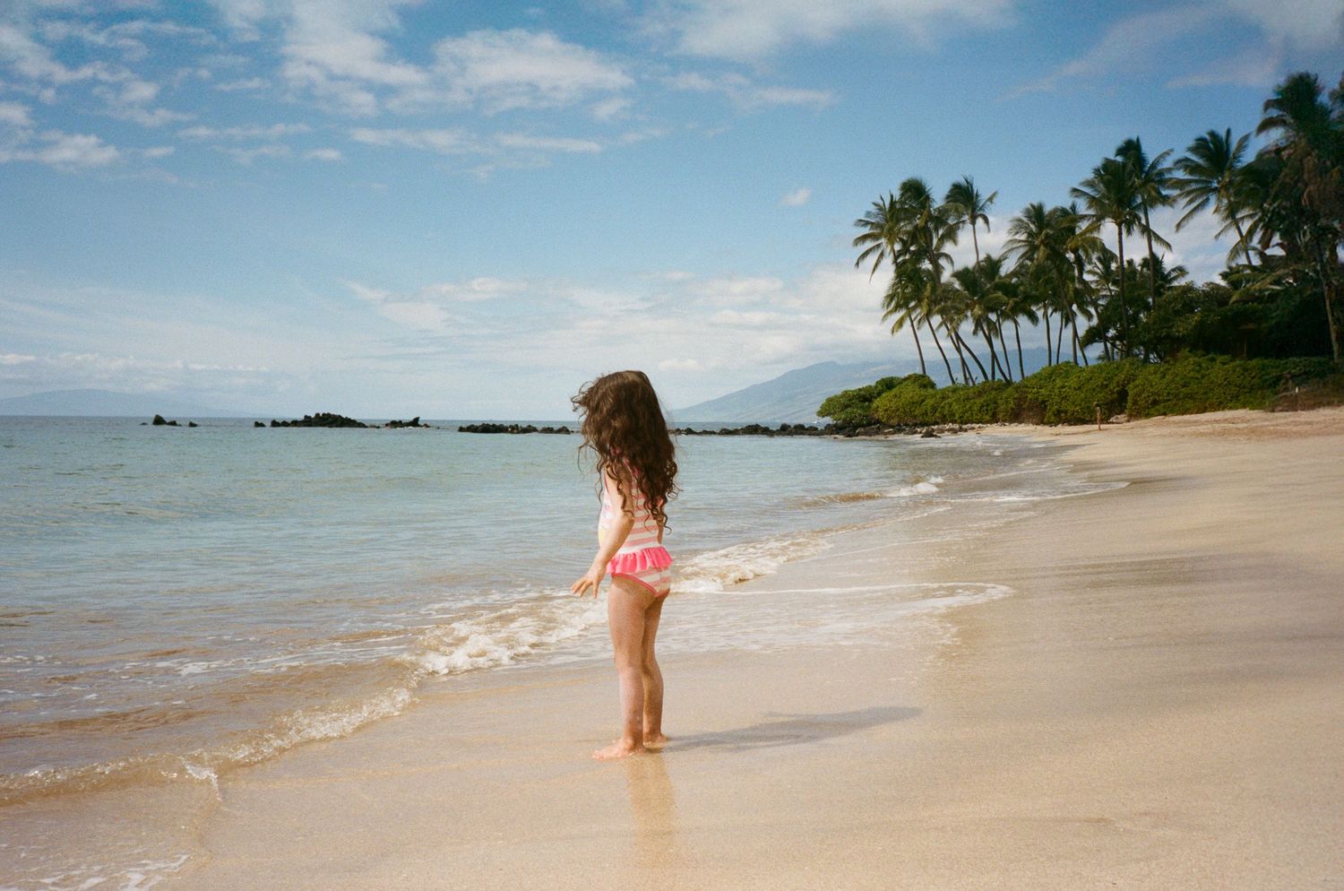 A person walks along a pristine sandy beach with palm trees and turquoise waters under a bright blue sky.