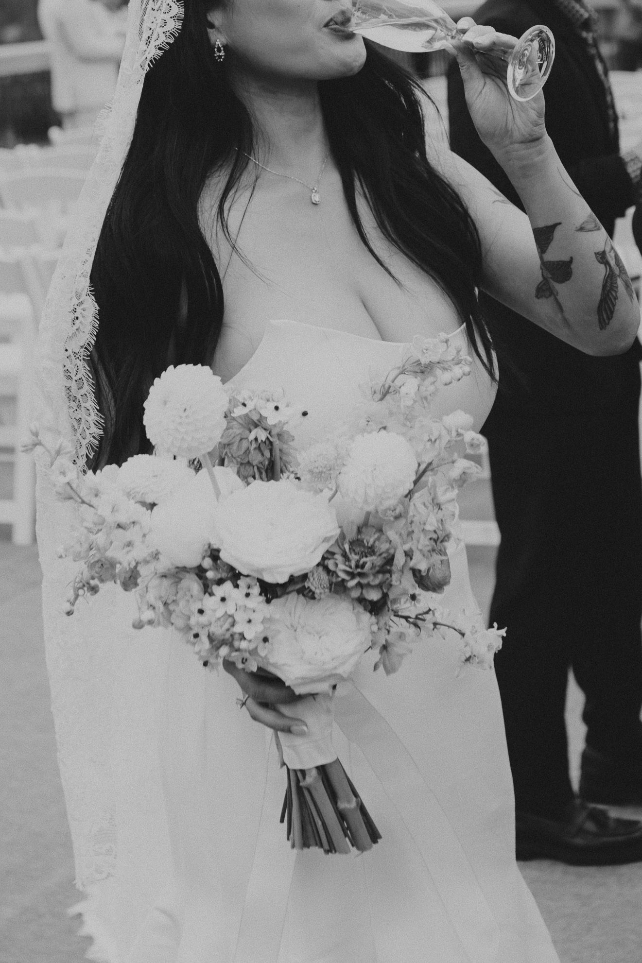 Elegant bride in lace dress holding a soft pastel bouquet while sipping champagne.