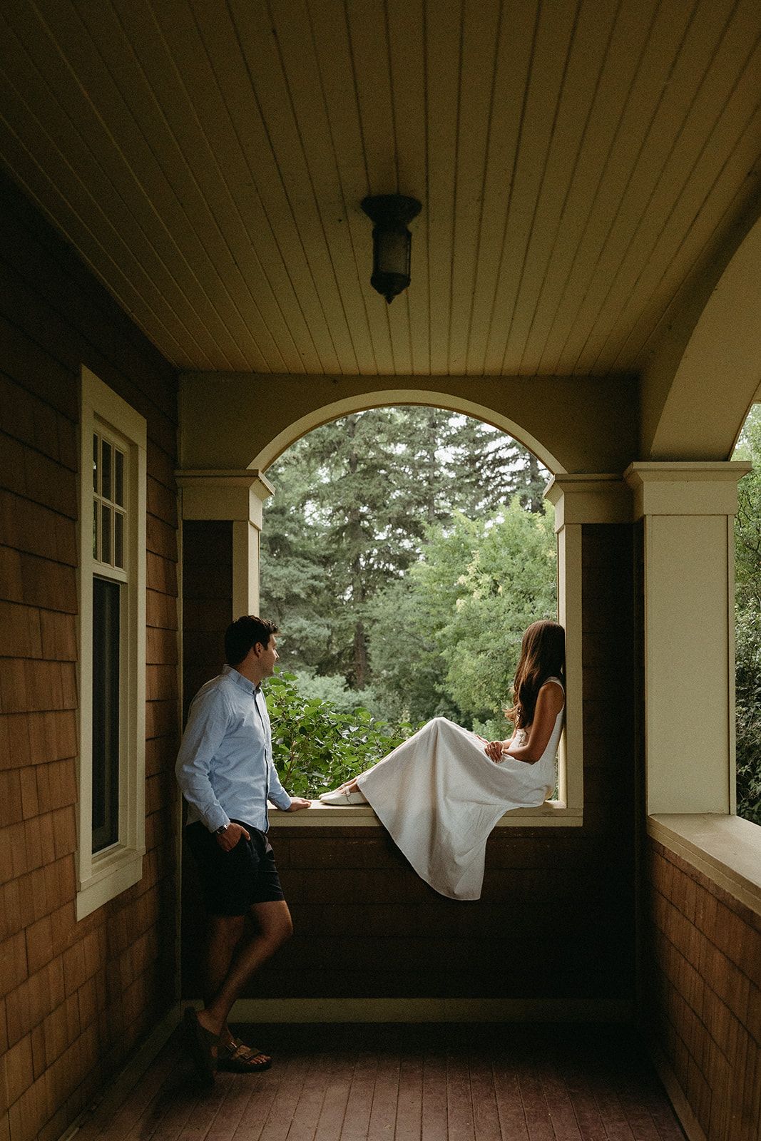 Couple sits in covered porch with wooden details and arched openings in moody lighting.