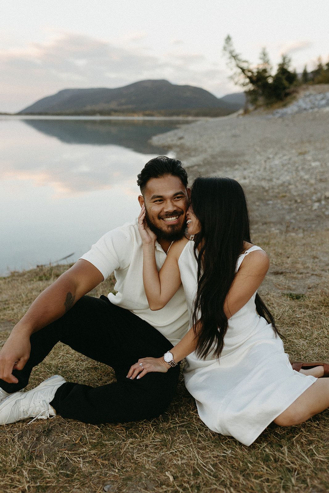 A couple shares intimate moments on a rocky beach with mountains in the background.