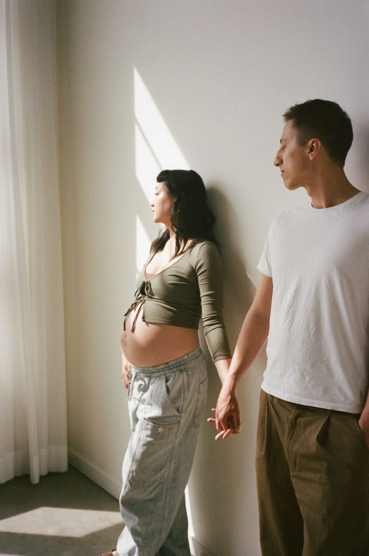 A couple stands near a window in a minimalist white room with soft natural lighting.