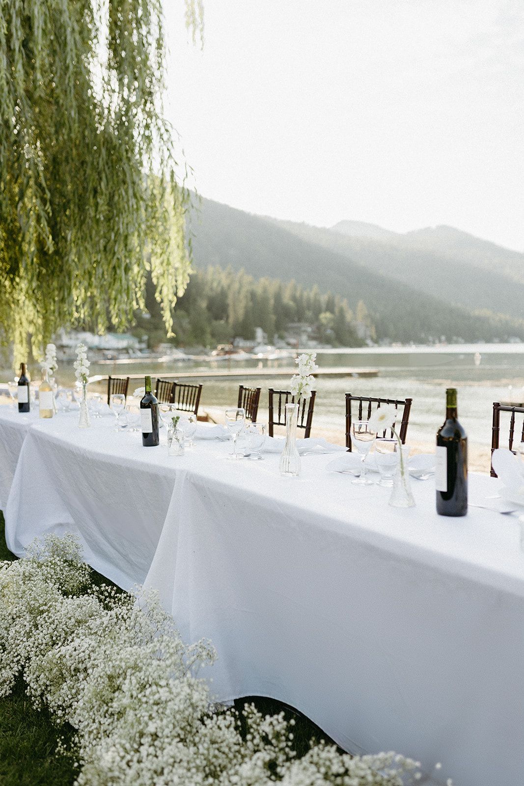 Long white reception tables set under weeping willow trees beside a mountain lake.