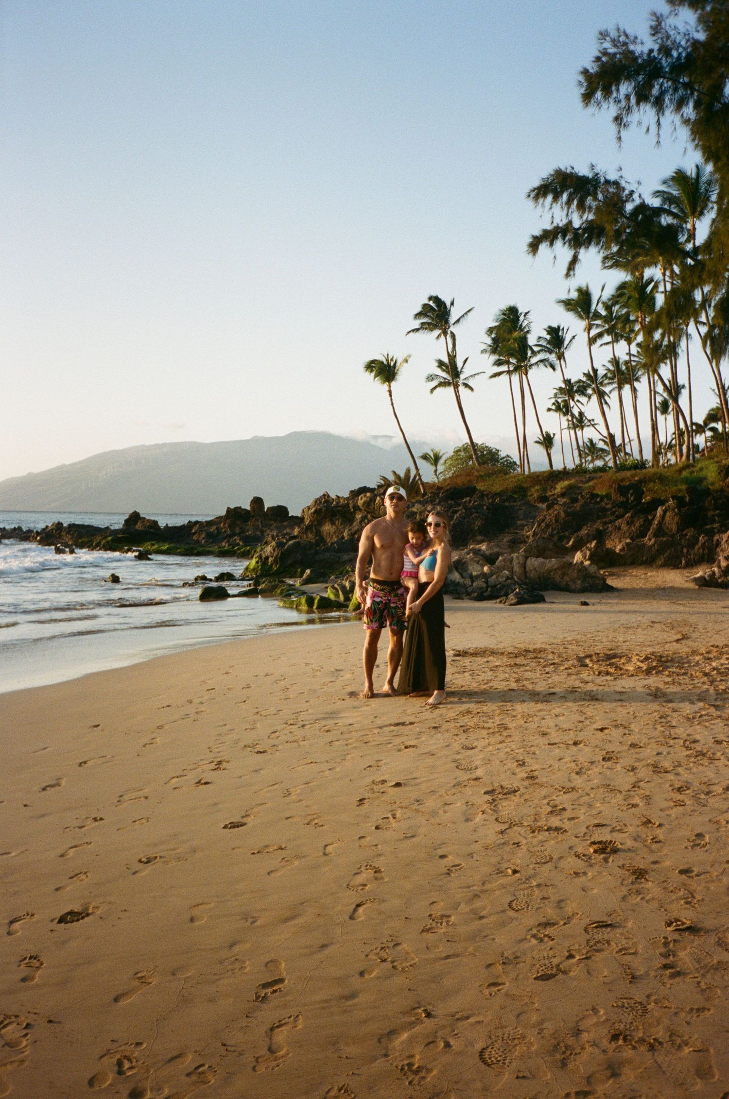 Palm trees sway in the breeze as beachgoers enjoy a scenic Hawaiian shoreline at sunset with mountains in the distance.