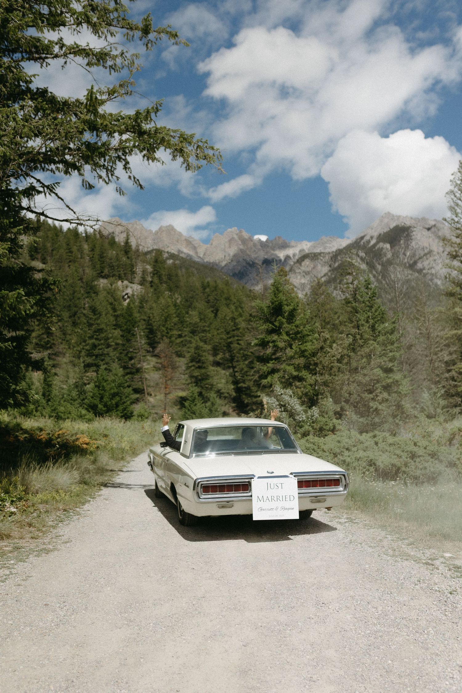 A vintage car sits parked on a mountain road with dramatic peaks and forest scenery in the background.