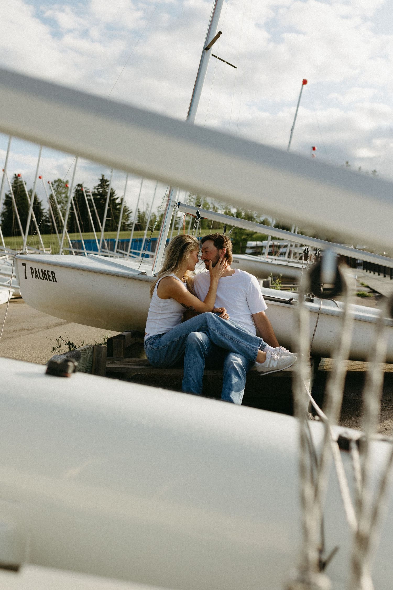 Couple sitting together on dock with sailboats and marina in background.