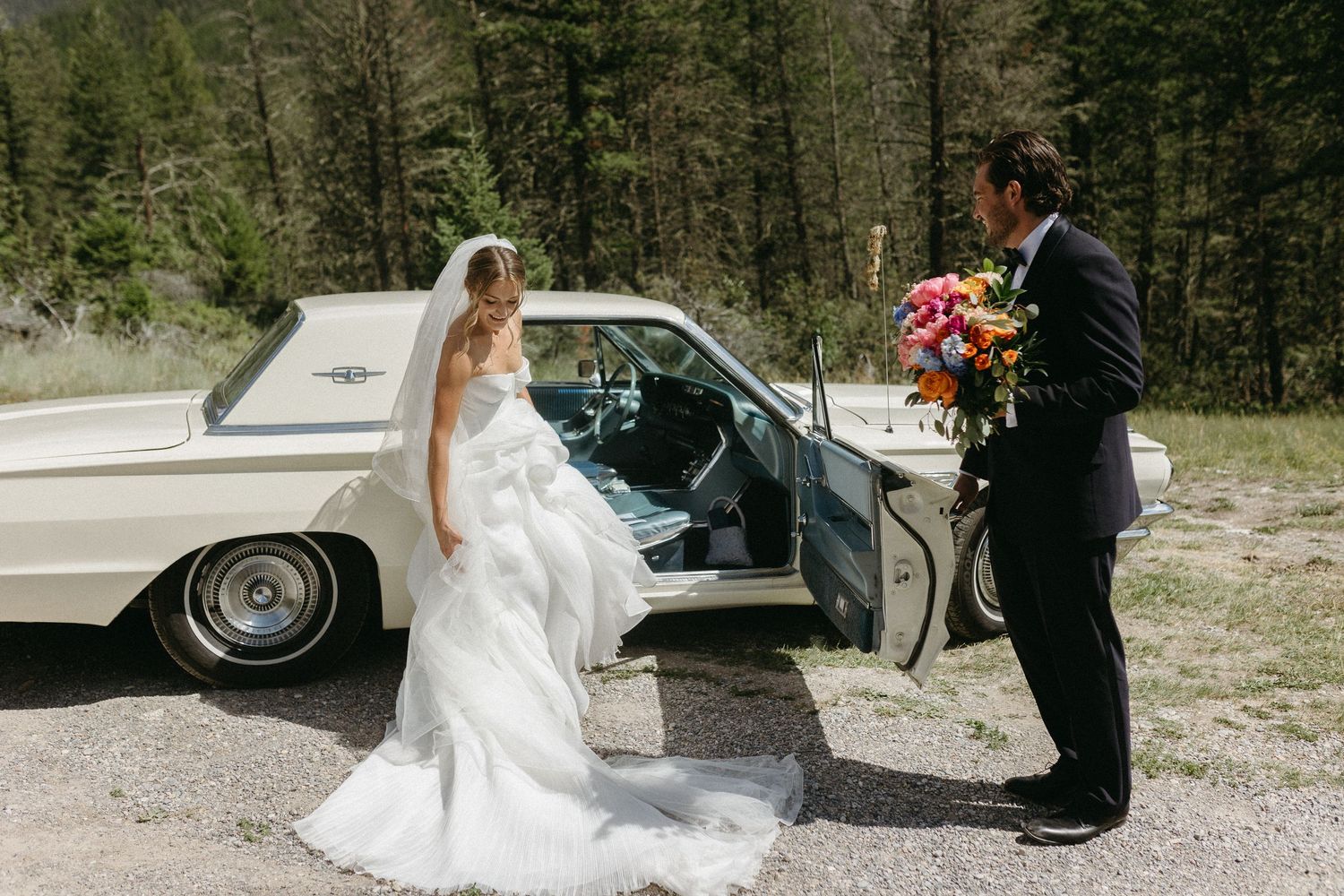 Wedding couple posing next to vintage car in mountain setting.