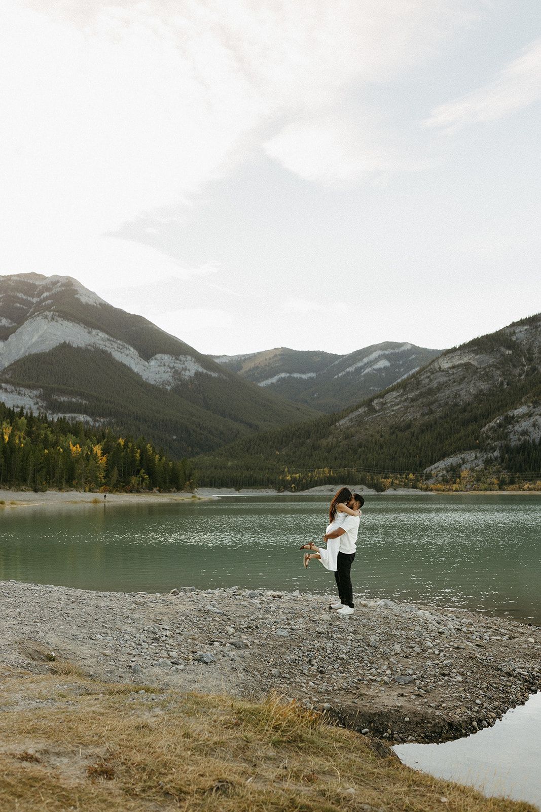 Couple embracing by a mountain lake with snow-capped peaks and evergreen forest in the background.