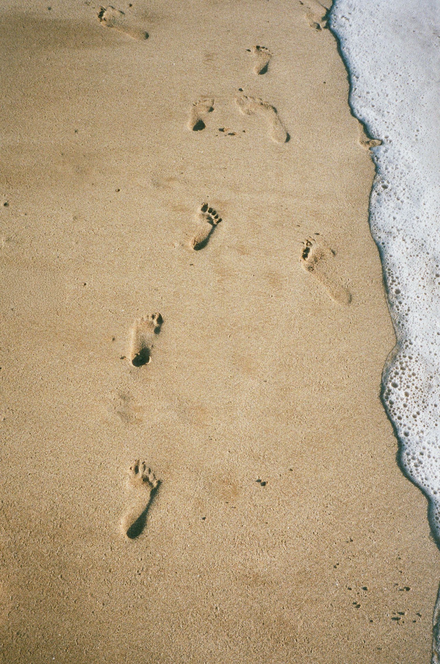 Footprints in wet sand along a foamy ocean shoreline create a peaceful beach scene.