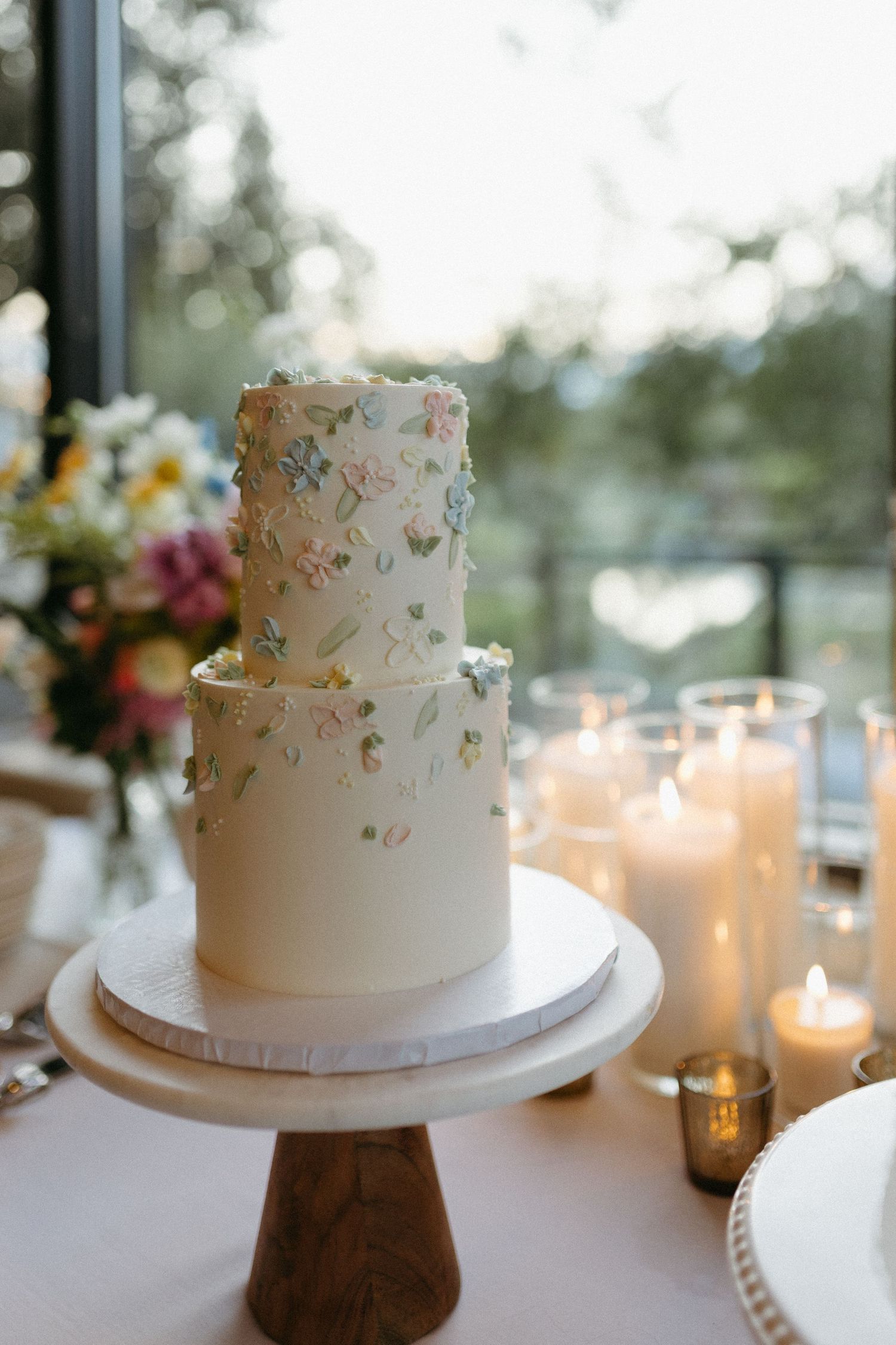 White wedding cake with delicate floral details displayed on wooden stand with glowing candles.