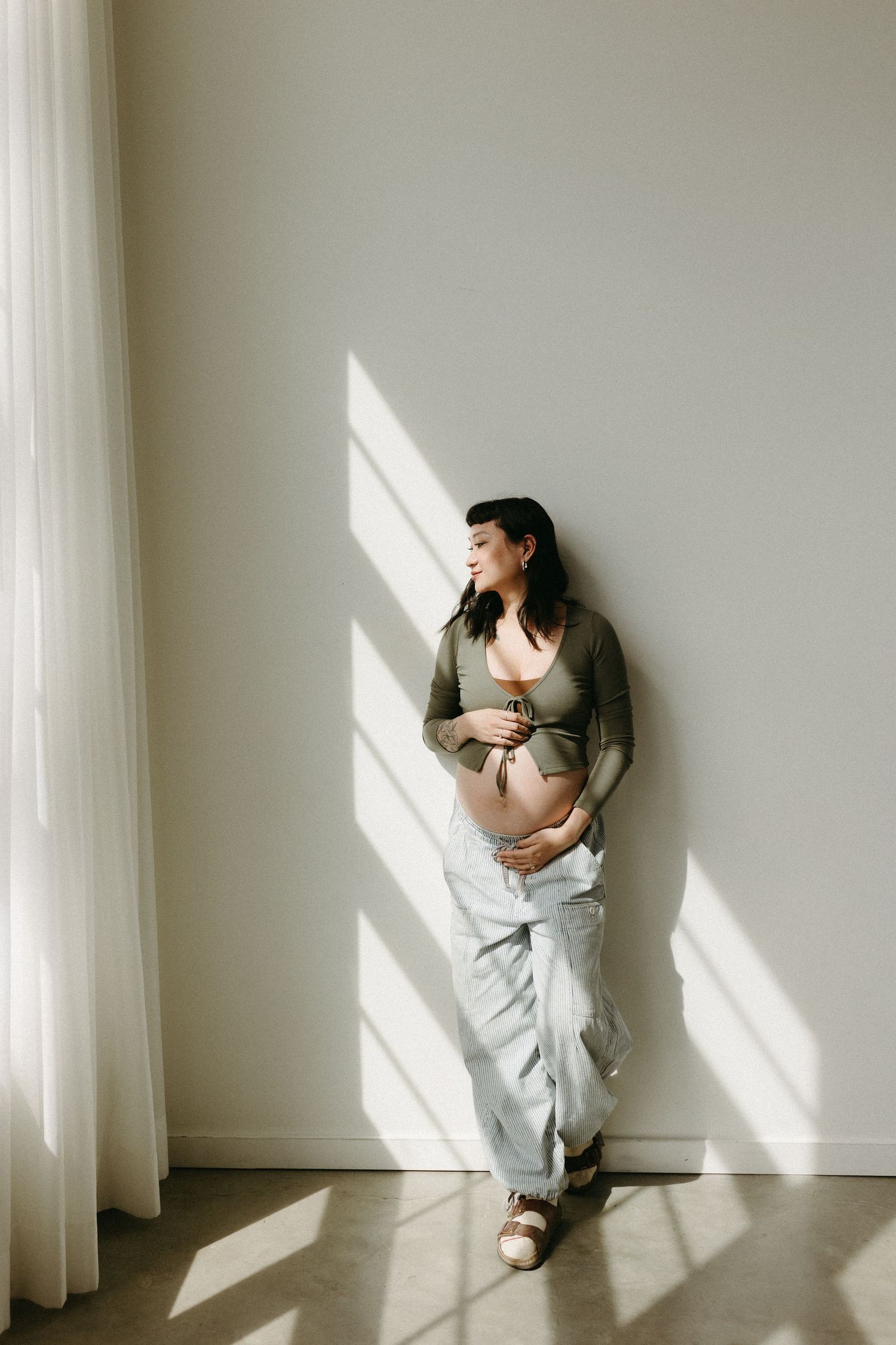 Artistic maternity portrait with dramatic window lighting on white wall.
