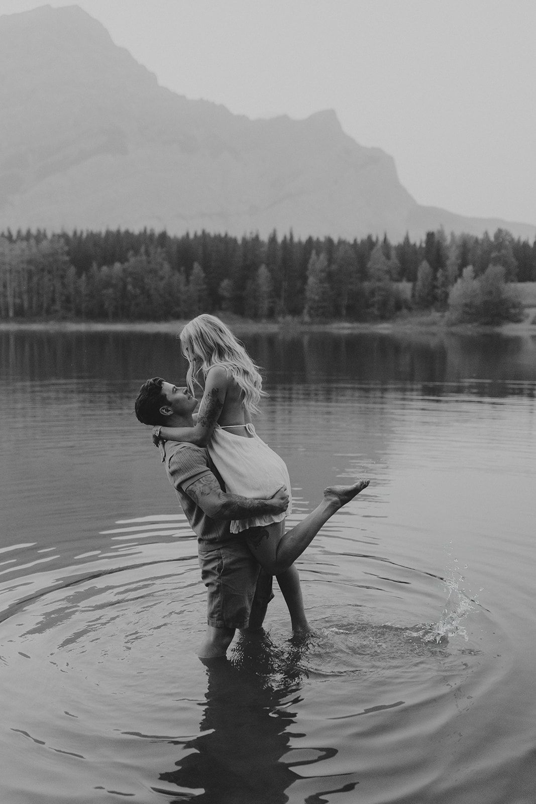 Black and white photo of a couple embracing in a mountain lake, man lifting woman with forest and peaks behind them.