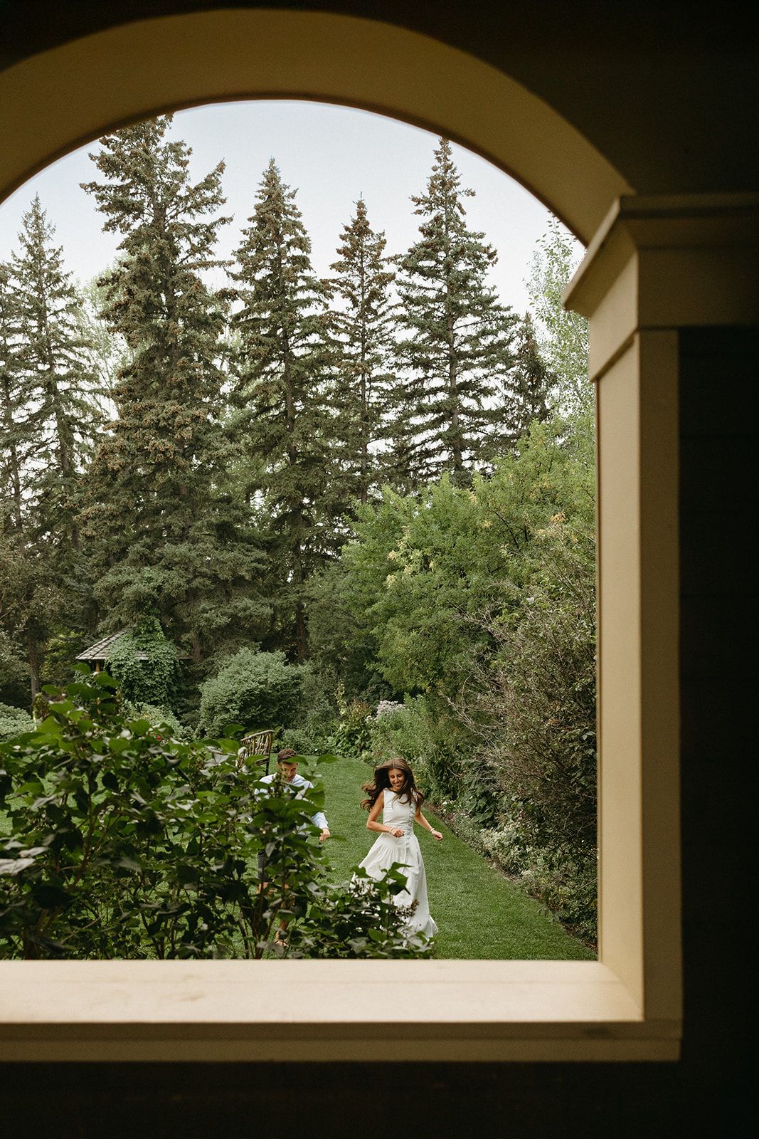 View through arched window frame of couple in garden with tall evergreen trees.