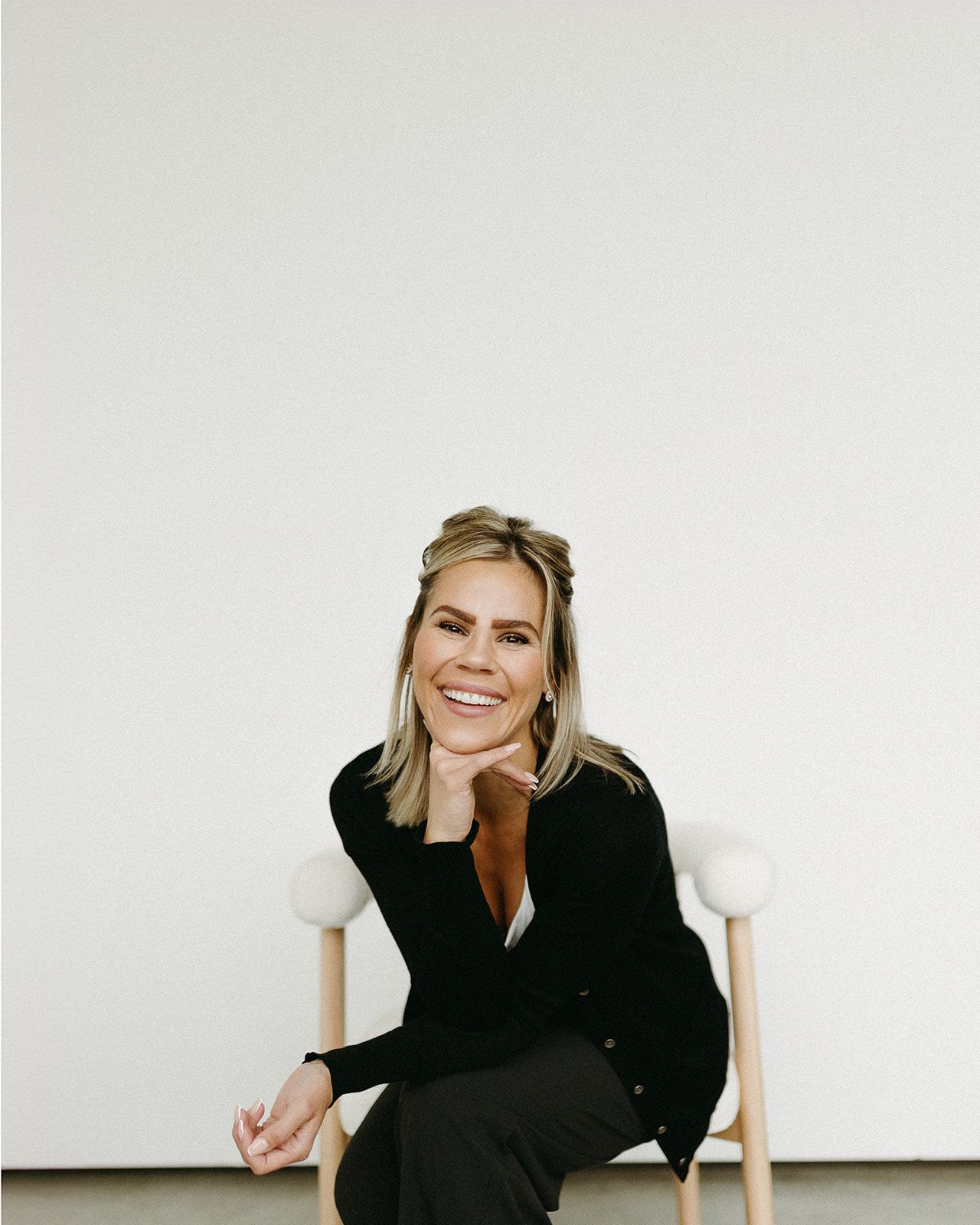 Person in black clothing sitting and laughing in a minimalist white setting.