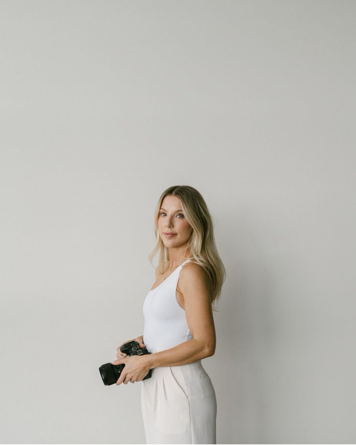 Woman in white sleeveless top and pants poses against light gray wall while holding sunglasses.