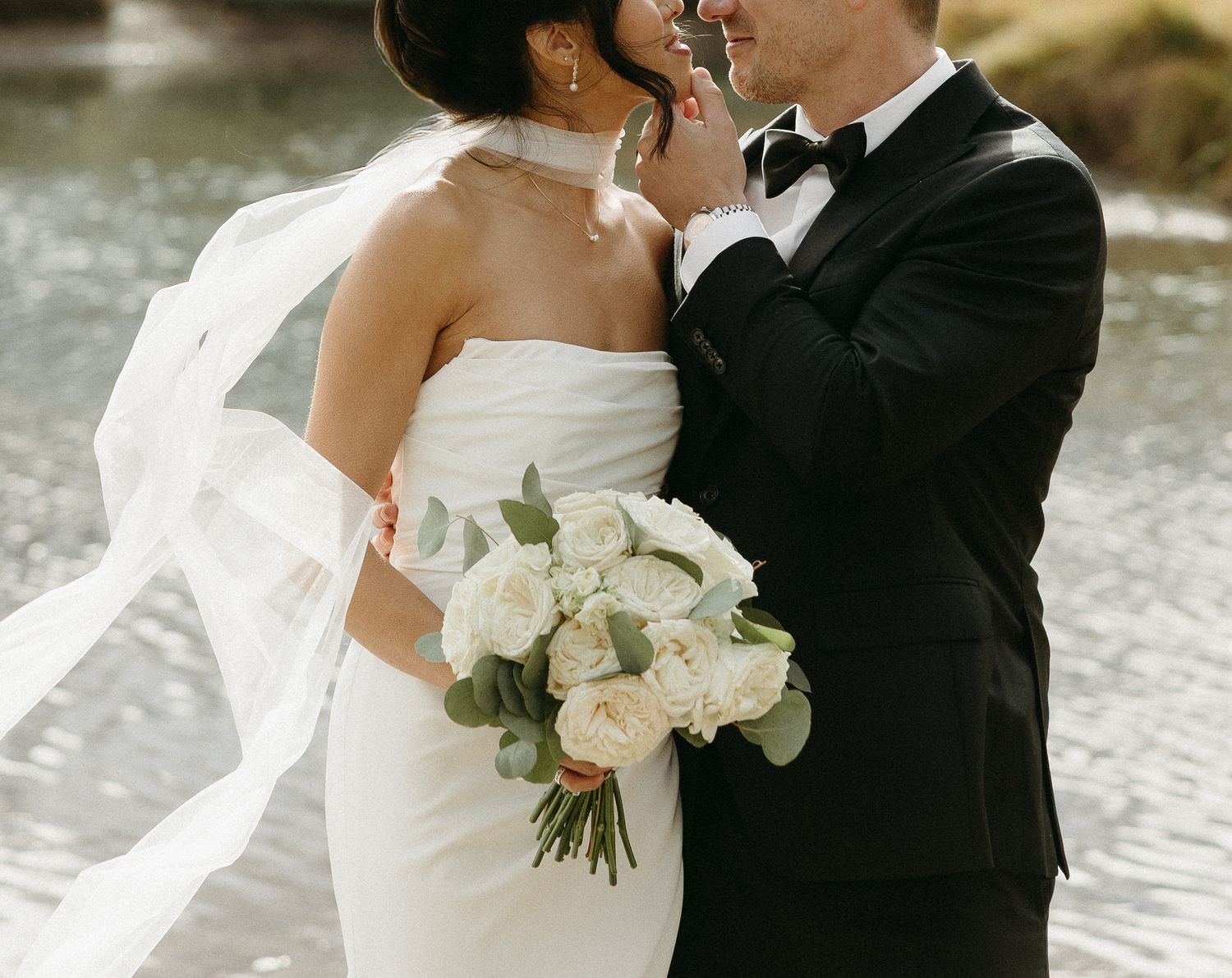 Romantic wedding couple embracing outdoors with white bouquet and flowing veil.