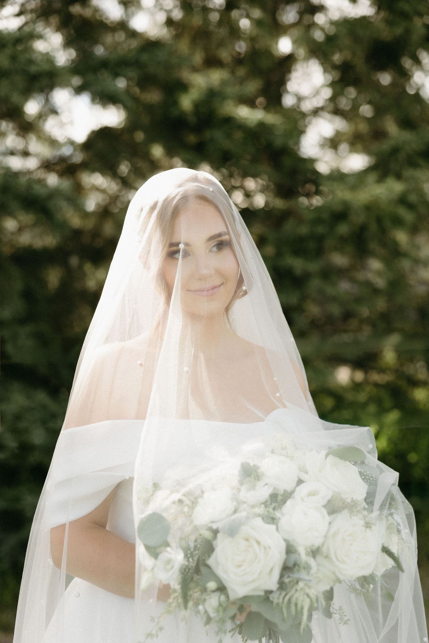 Ethereal bridal portrait with white roses bouquet through a sheer veil outdoors.
