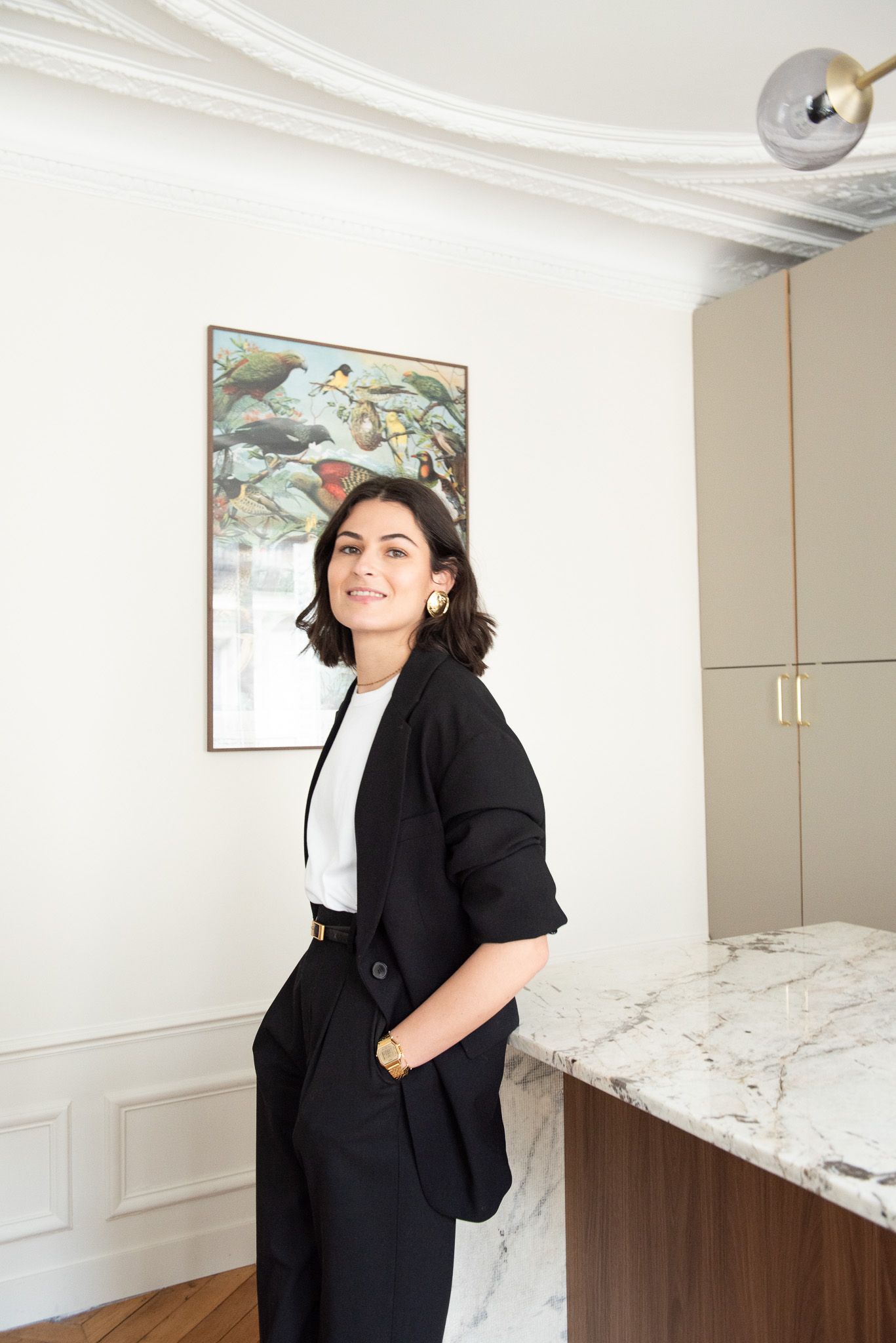 Woman in black blazer smiling beside marble kitchen island in elegant Parisian apartment.