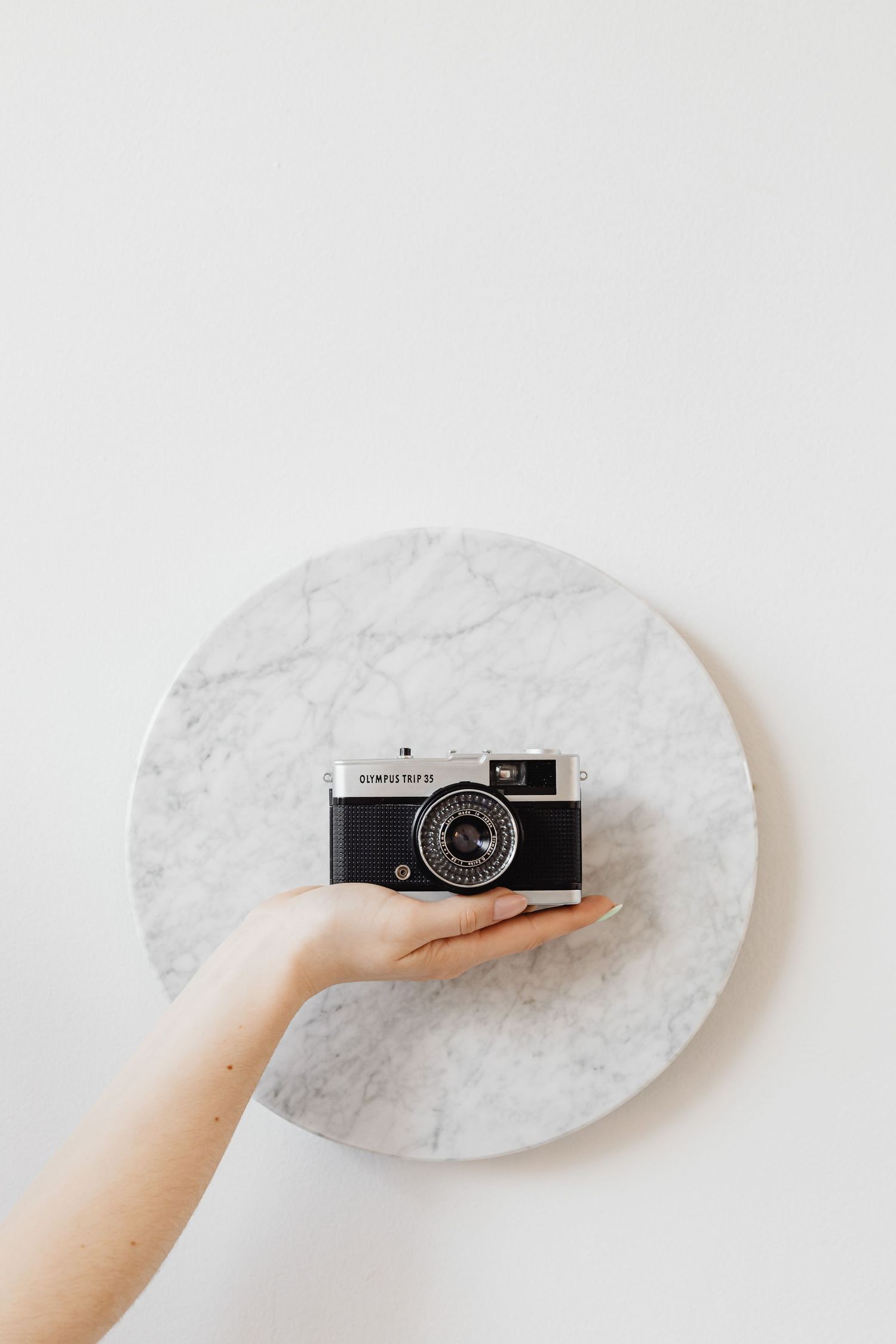 Hand holding a vintage Olympus Trip 35 film camera against a round marble surface on white background.