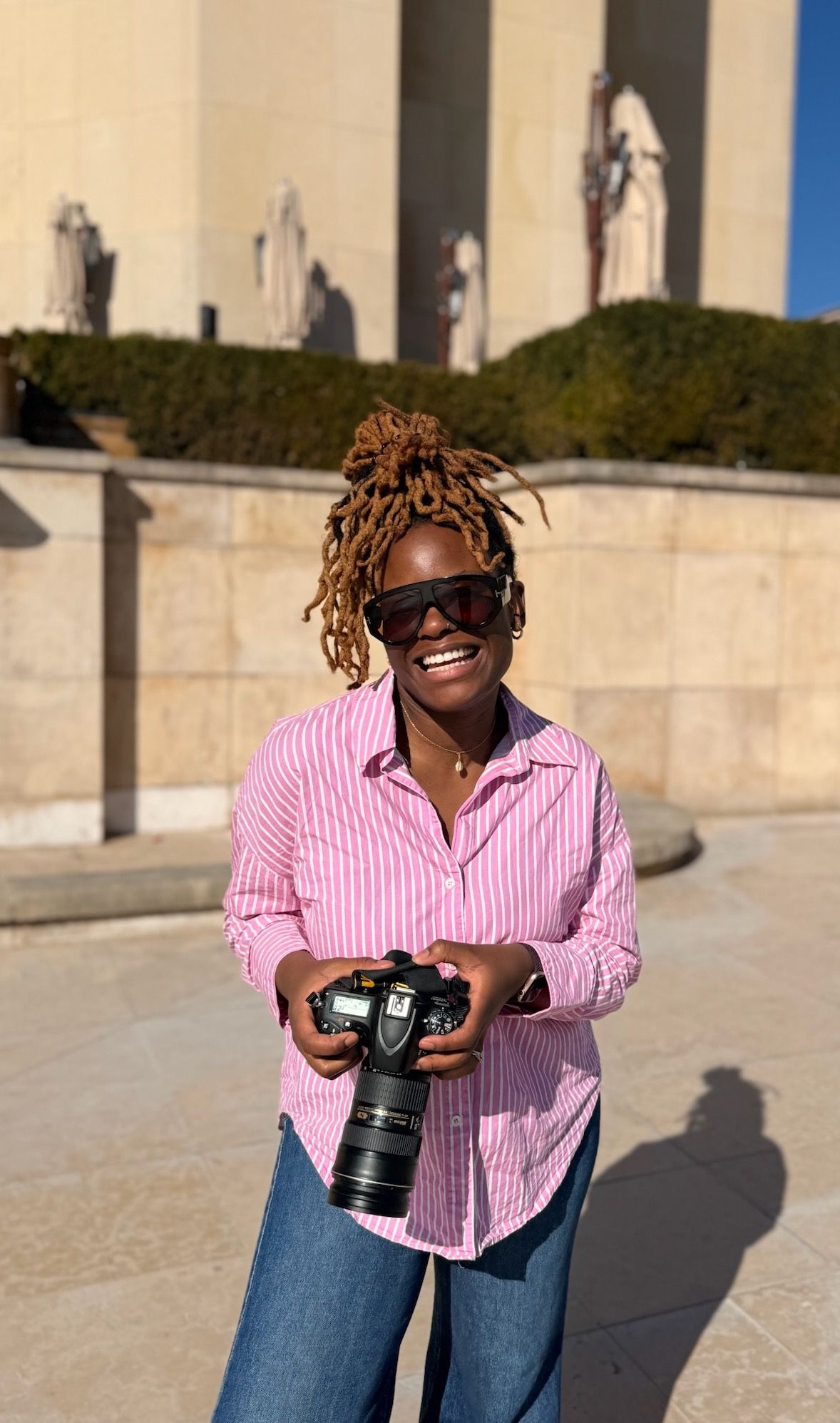 Femme souriante aux cheveux locksés, tenant un appareil photo reflex numérique, vêtue d'une chemise à rayures roses et de lunettes de soleil