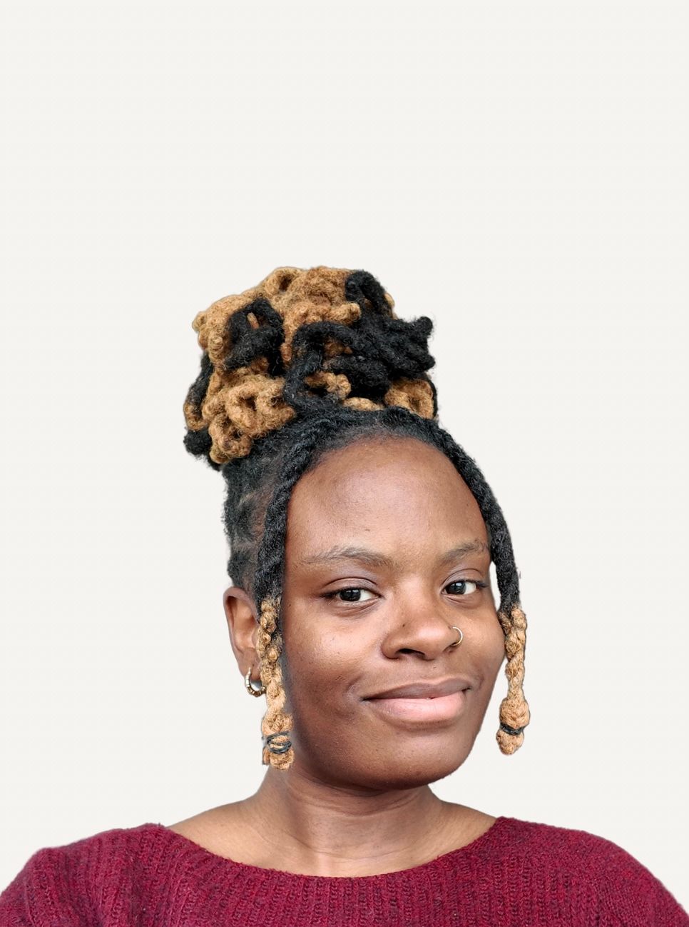 Woman with styled locs updo and gold earrings wearing a burgundy sweater, smiling against a light background.