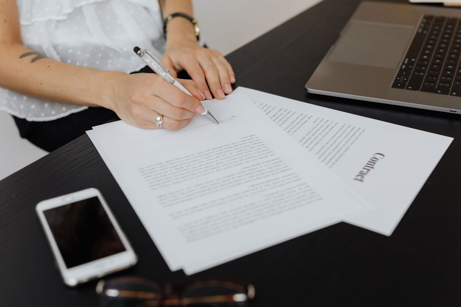 Une personne signe un contrat à un bureau, avec un ordinateur portable un Macbook Pro, un téléphone iPhone et des lunettes à proximité.