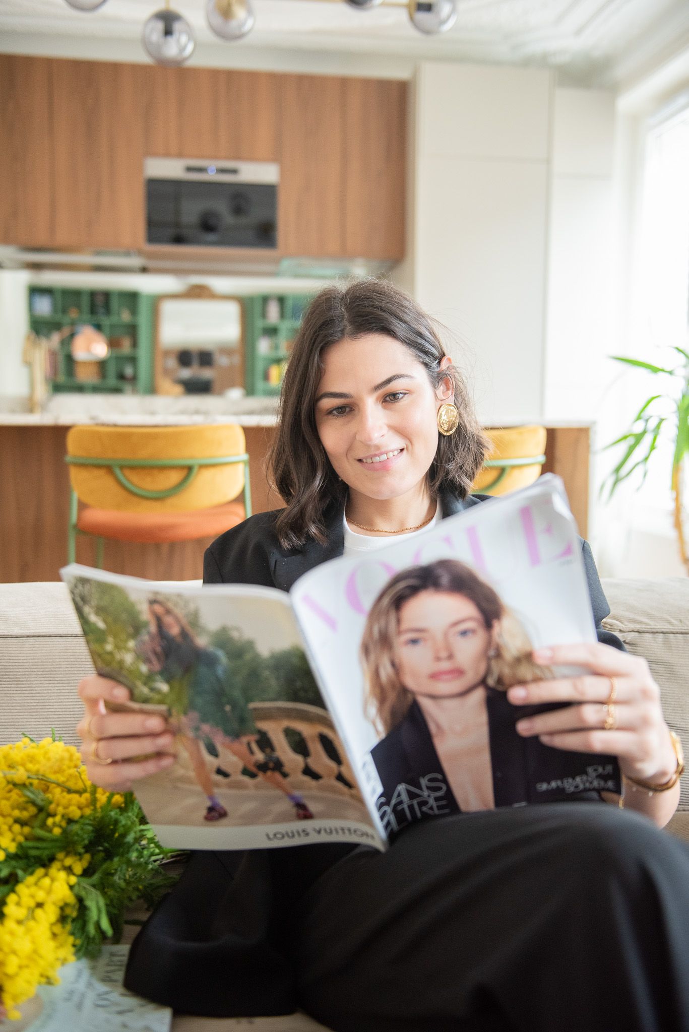 Woman in black outfit reading Vogue magazine on sofa in stylish apartment with wood kitchen.
