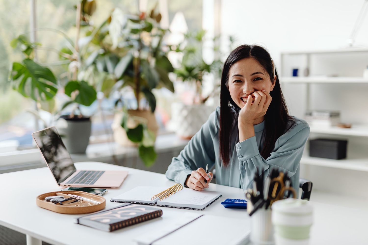 Smiling Asian woman sitting at desk with laptop and notebook, surrounded by green plants in bright home office.