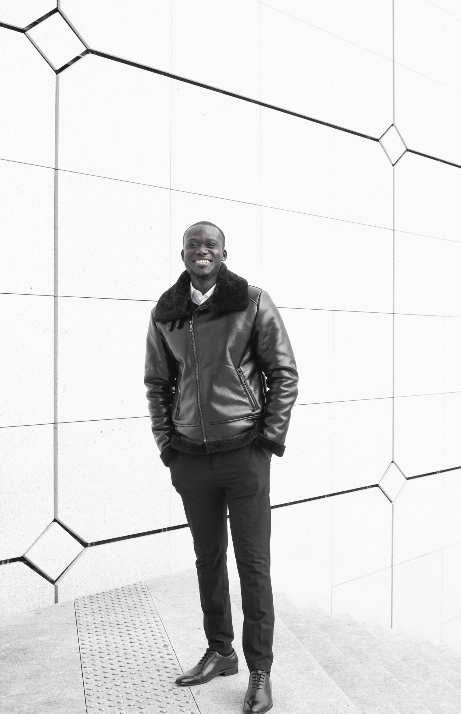Smiling Black man in leather jacket posing confidently against a geometric patterned white wall in black and white.