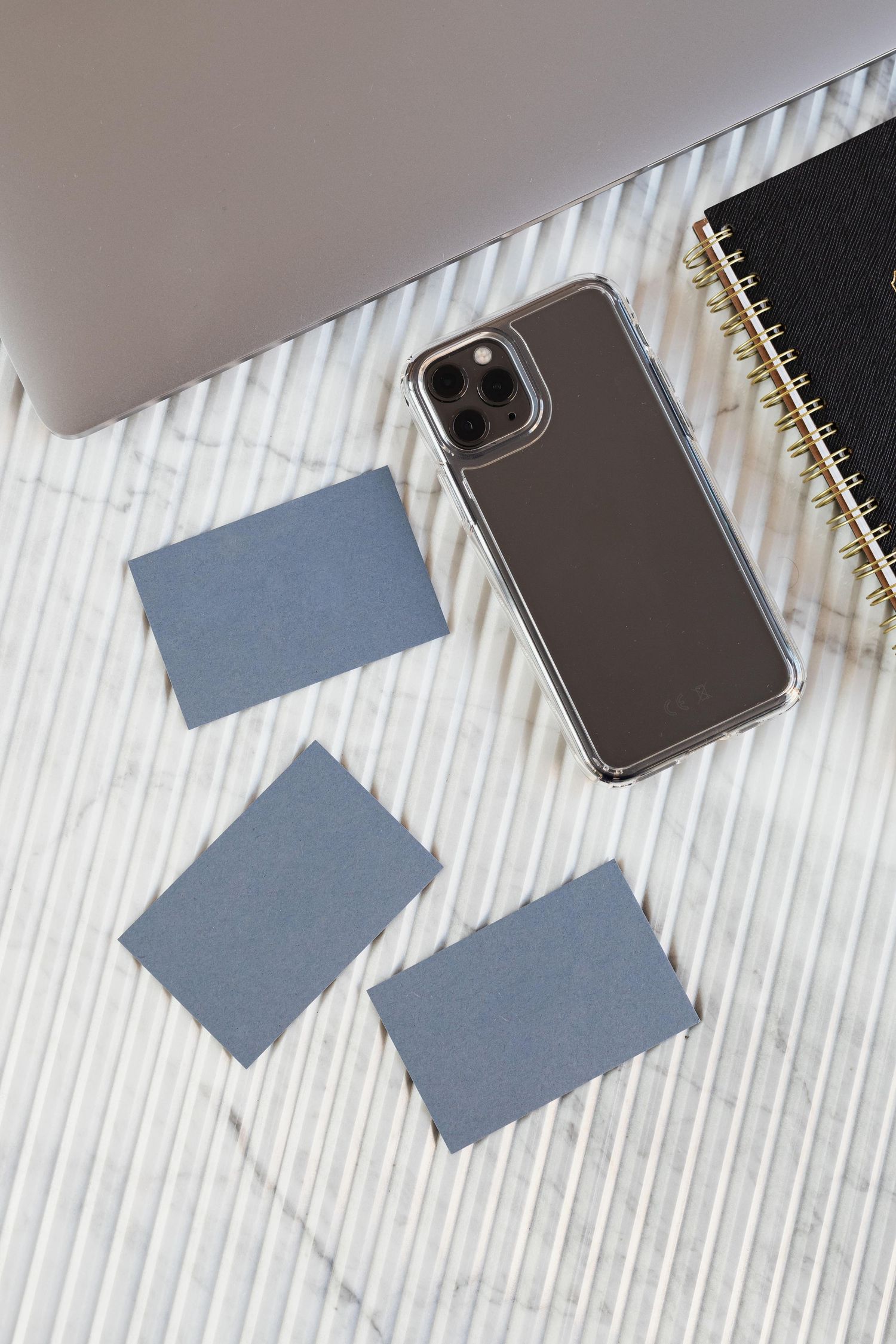Flat lay of gray business cards, smartphone, spiral notebook, and laptop on marble surface.
