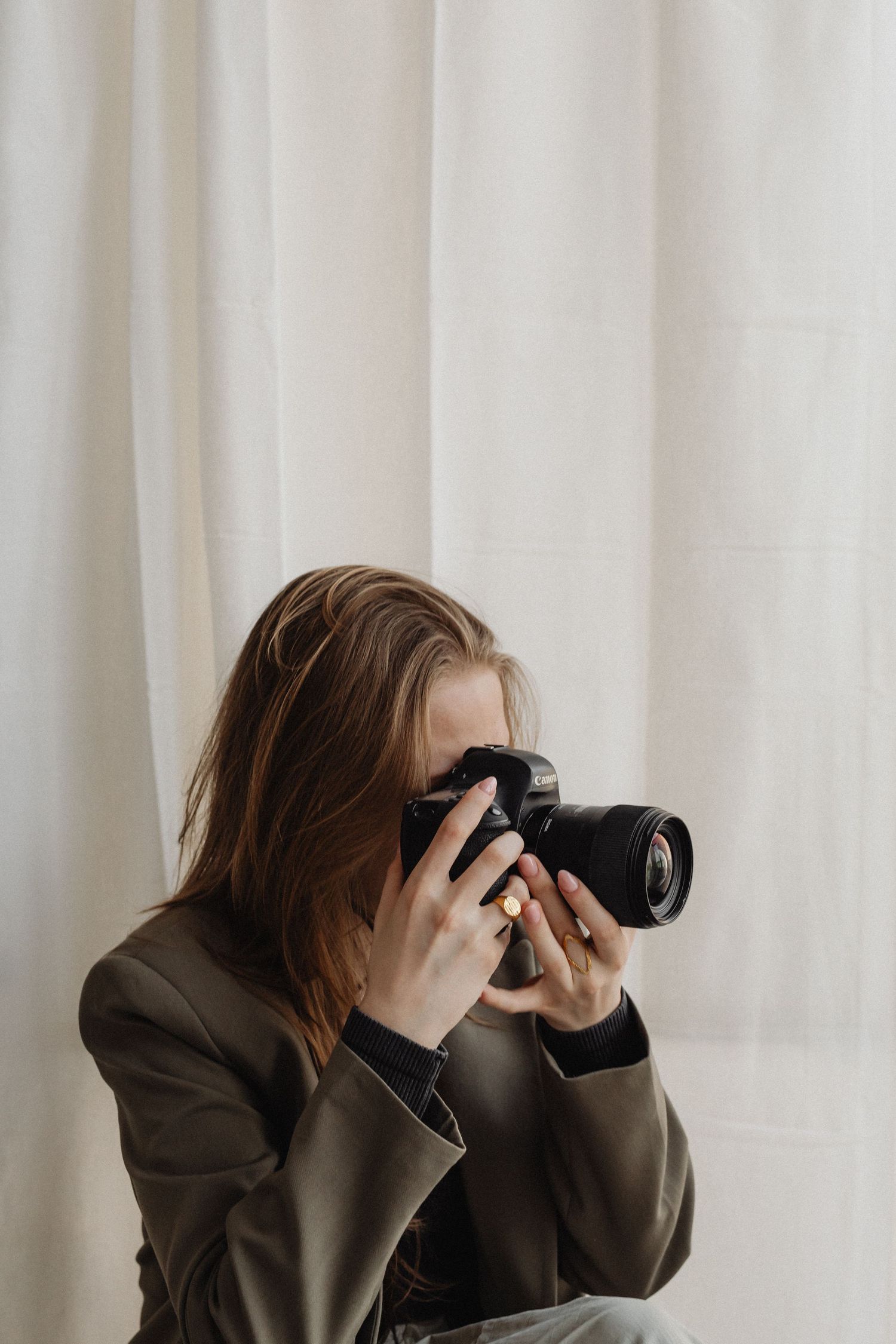 Young woman with brown hair photographing with DSLR camera in front of white linen curtain.