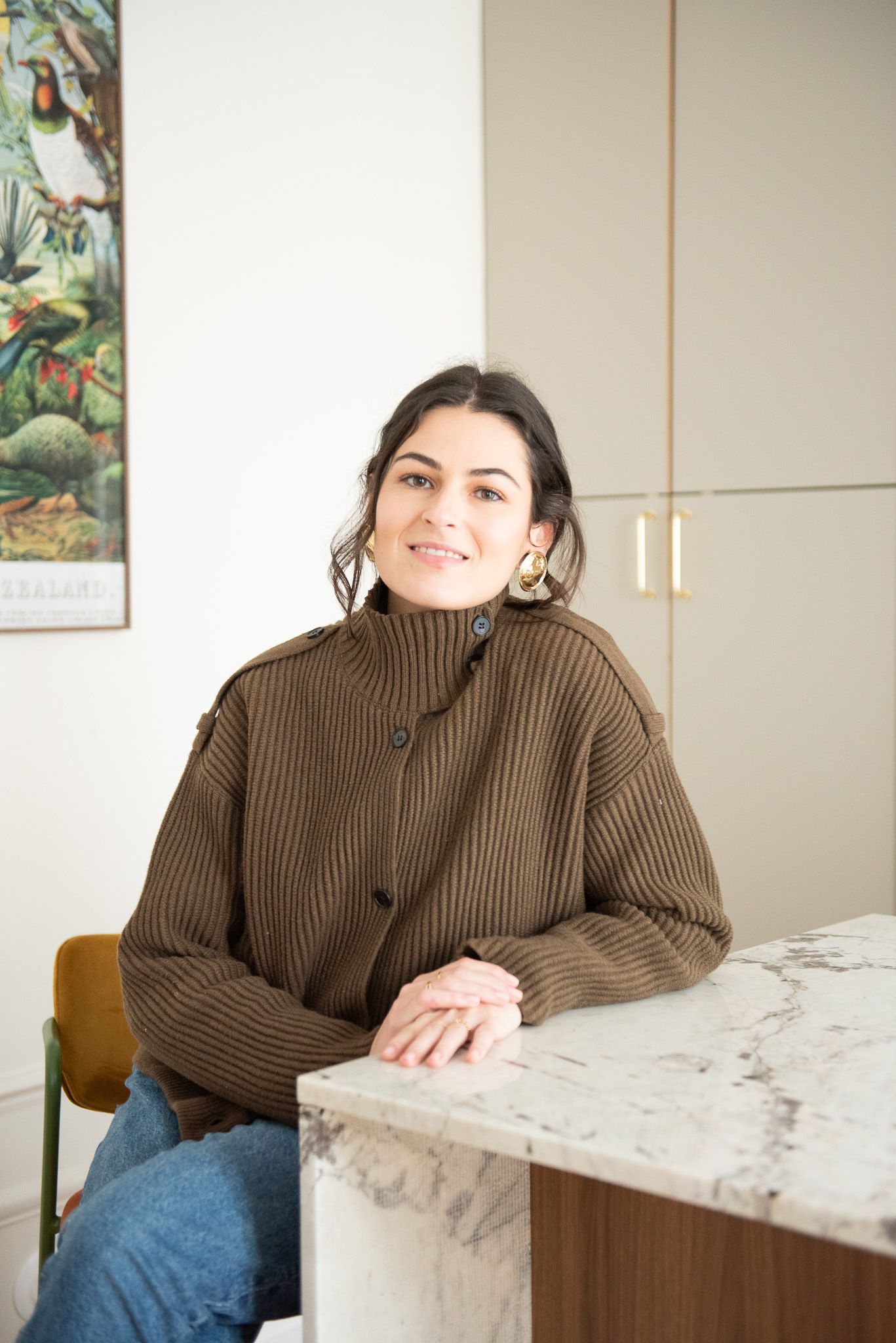 Woman in brown ribbed sweater smiling at marble counter, gold hoop earrings, nature poster on white wall behind her.