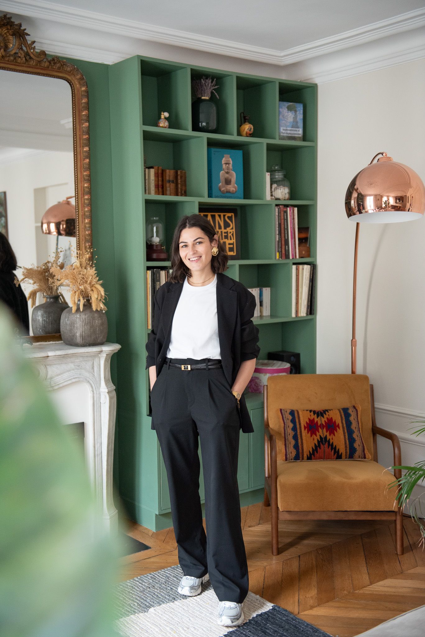 Smiling woman in black blazer and white tee standing in living room with green bookshelf.